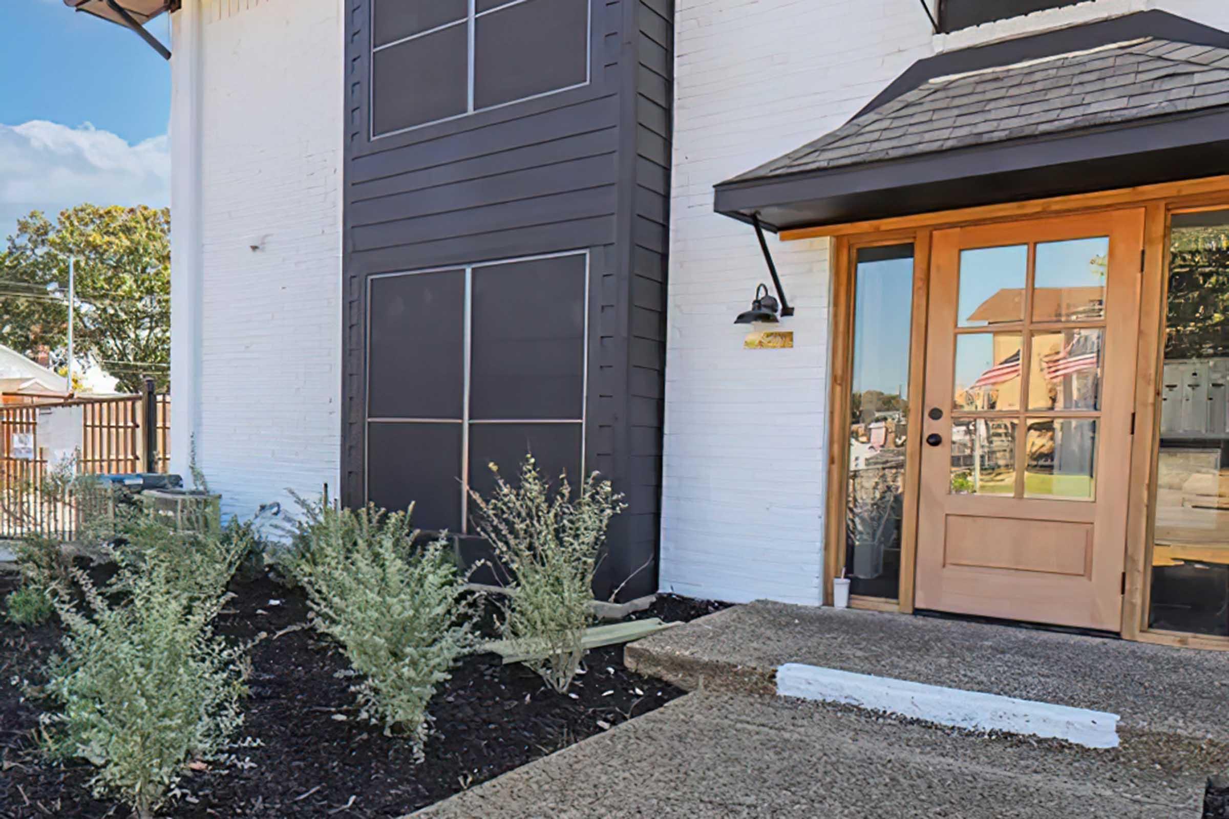 A welcoming entrance featuring a wooden door with a decorative awning, flanked by large windows. The pathway is made of textured concrete, leading to a well-maintained garden area with green plants. In the background, a hint of a flag can be seen, adding a touch of charm to the scene.