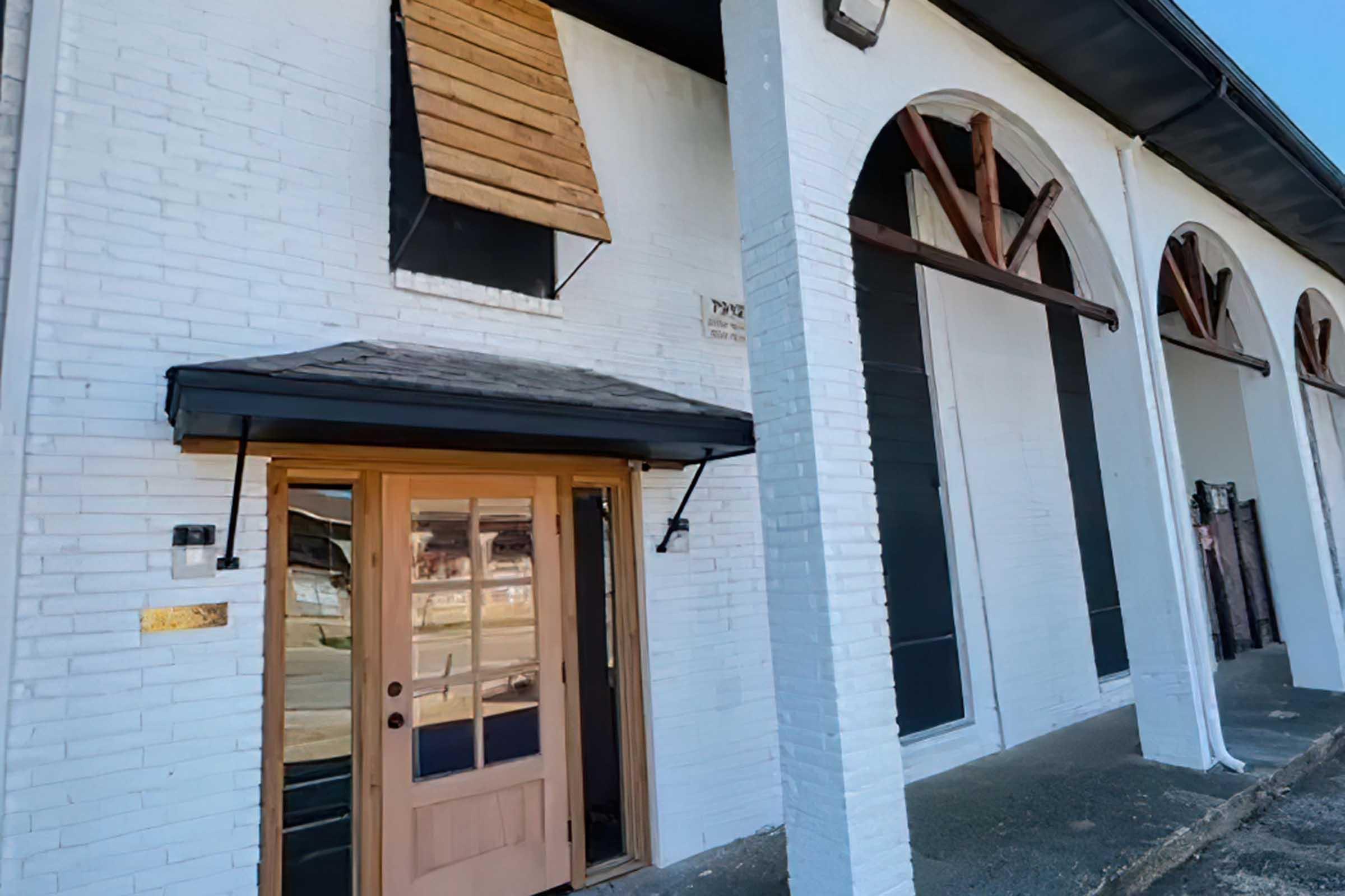 Exterior view of a white brick building featuring a wooden door with a transom window above it. There are several closed windows with wooden shutters and an arched design above some of the windows. The setting appears to be a quiet street with limited activity.