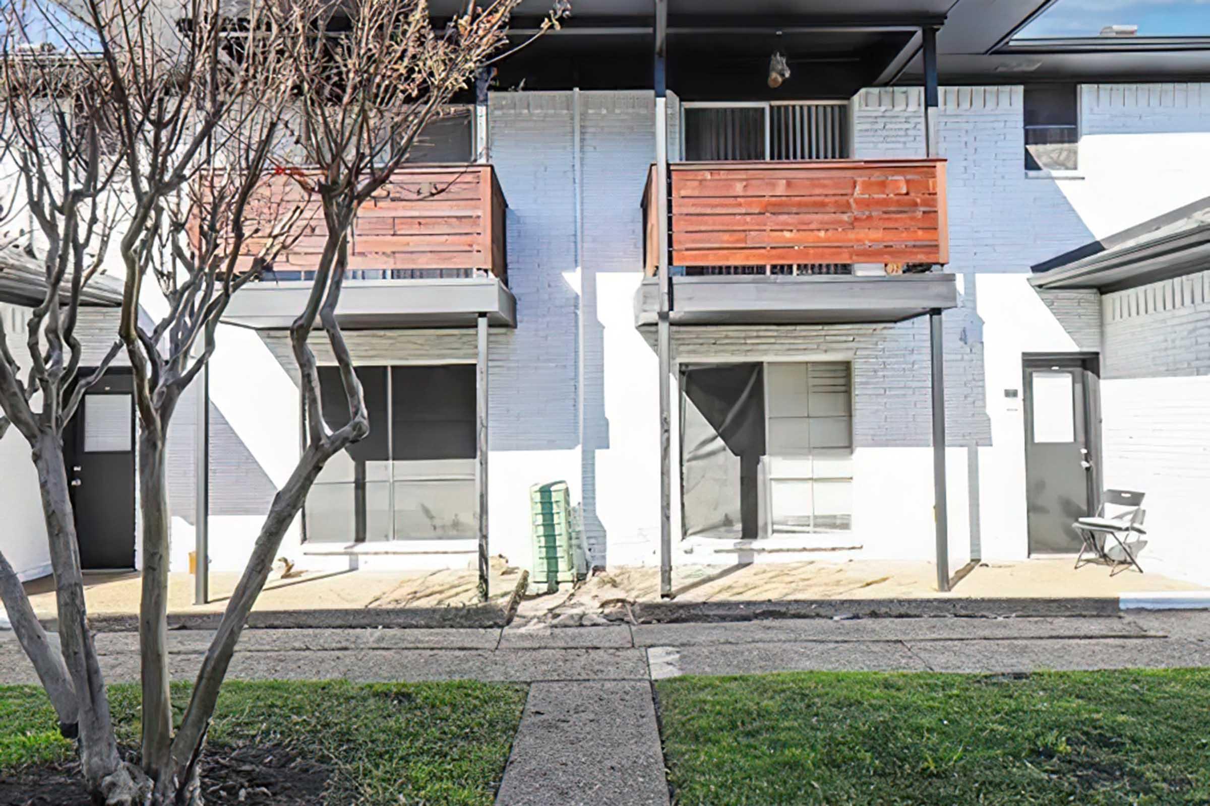 Two-story building exterior featuring two balconies with wooden railings. The ground level has entrances with glass doors, and a small patch of green grass is visible in front of the building. There is a bare tree and a green storage unit on the ground. The overall environment appears tidy and well-maintained.