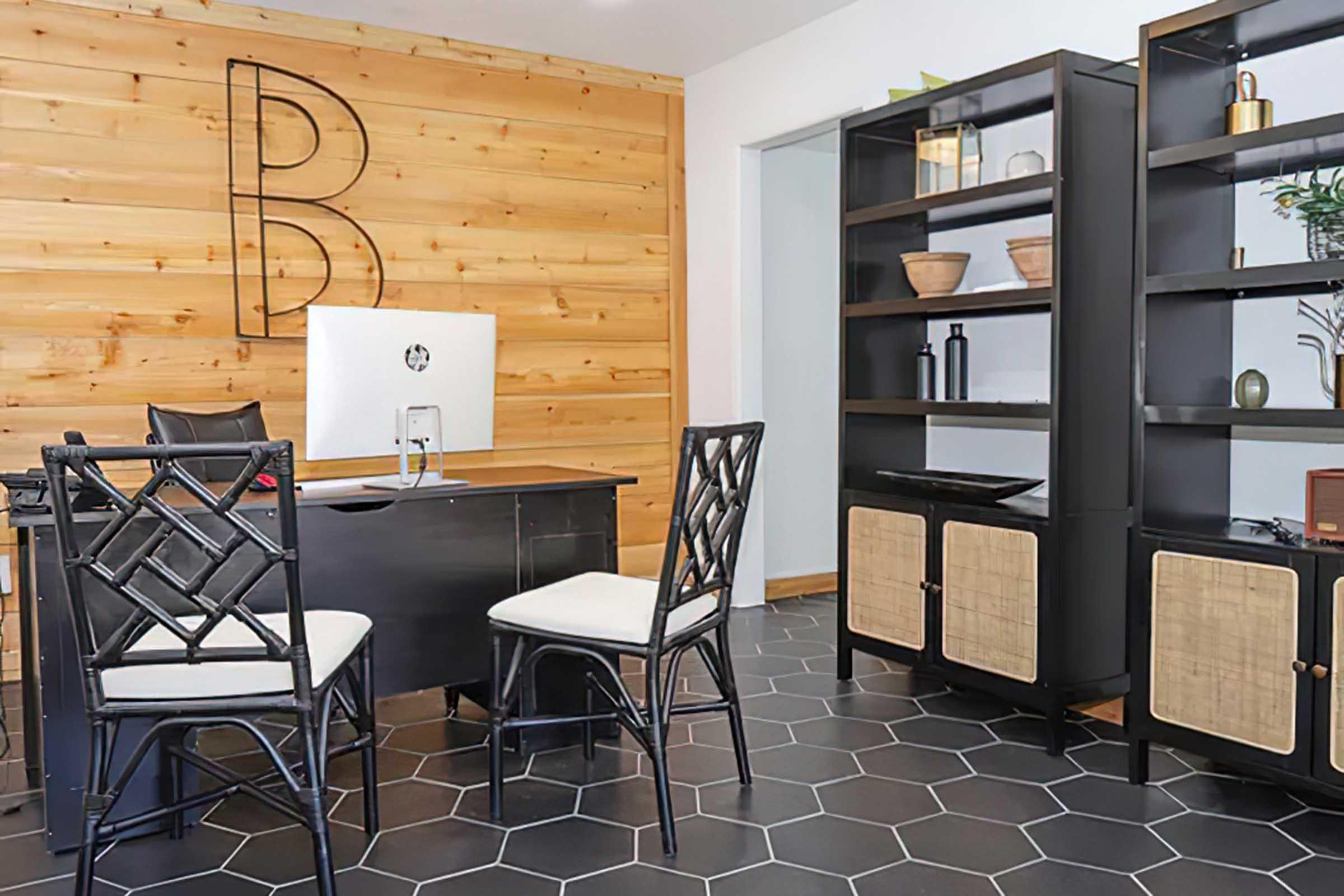 A modern office space featuring a black desk with an iMac, two stylish chairs, and a shelving unit. The wall behind is adorned with wooden panels and a letter "B." The floor is hexagon-patterned tiles, creating a contemporary and inviting atmosphere.
