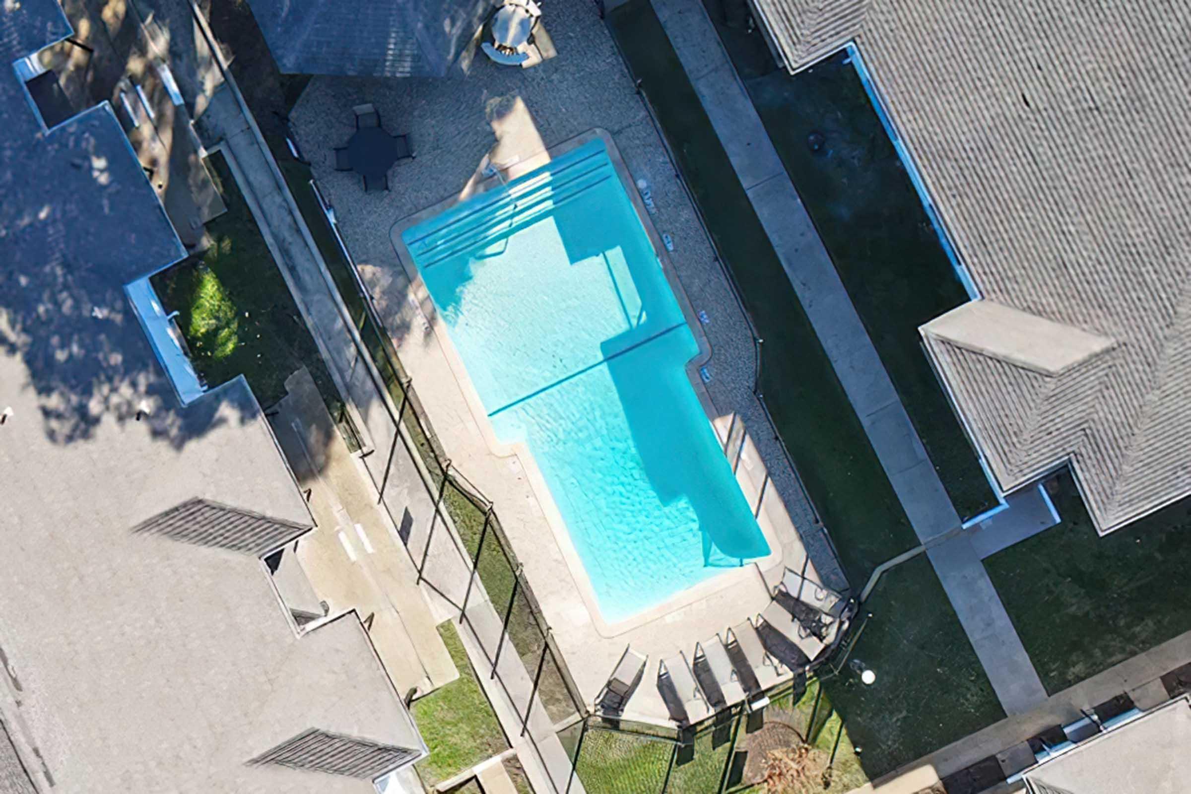Aerial view of a swimming pool surrounded by a fence, with lounge chairs positioned nearby. The pool is clear and rectangular, situated between two residential buildings. Sunlight casts shadows on the pool area, while green grass is visible around the perimeter.