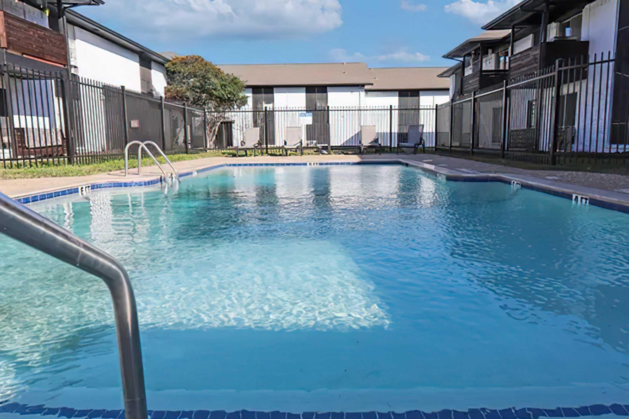 A clear swimming pool surrounded by a metal fence, with lounge chairs and buildings visible in the background. The water reflects the blue sky and clouds above, creating a serene and inviting atmosphere.