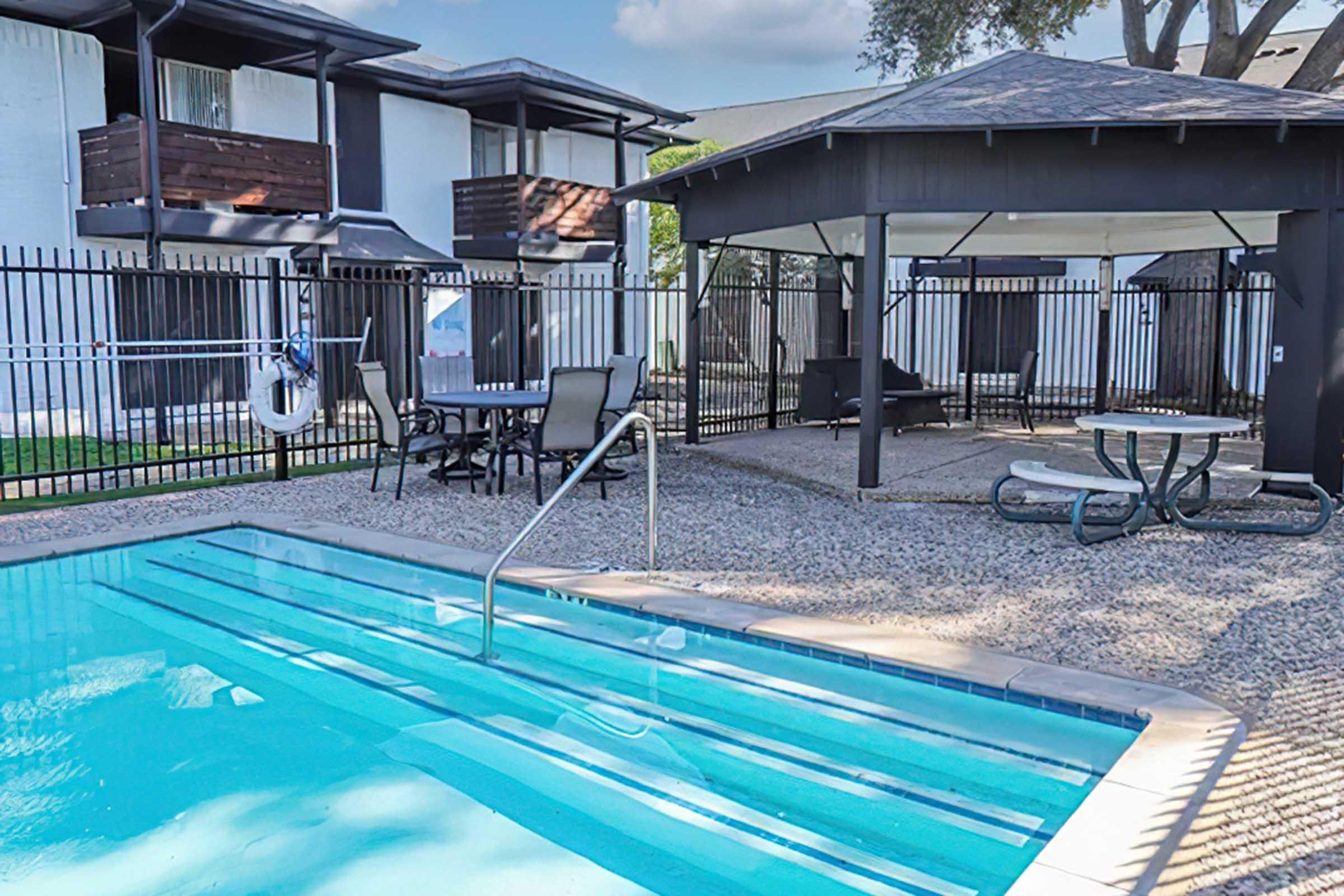 A clear swimming pool with a ladder, surrounded by a gravel area. In the background, there are two covered seating areas and an outdoor dining set. The pool area is enclosed with a black fence, and nearby structures suggest a residential or apartment complex.