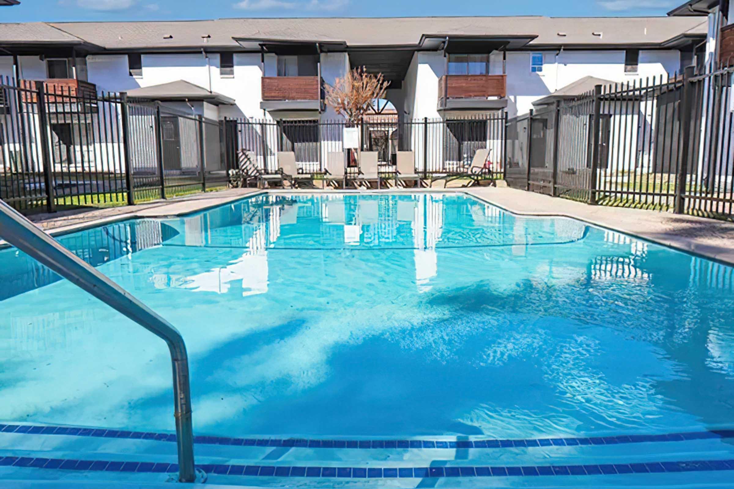 A clear blue swimming pool surrounded by a fence, with lounge chairs positioned nearby. In the background, there are apartments with balconies and a tree. The scene is bright and inviting, conveying a relaxing atmosphere typical of a residential complex.