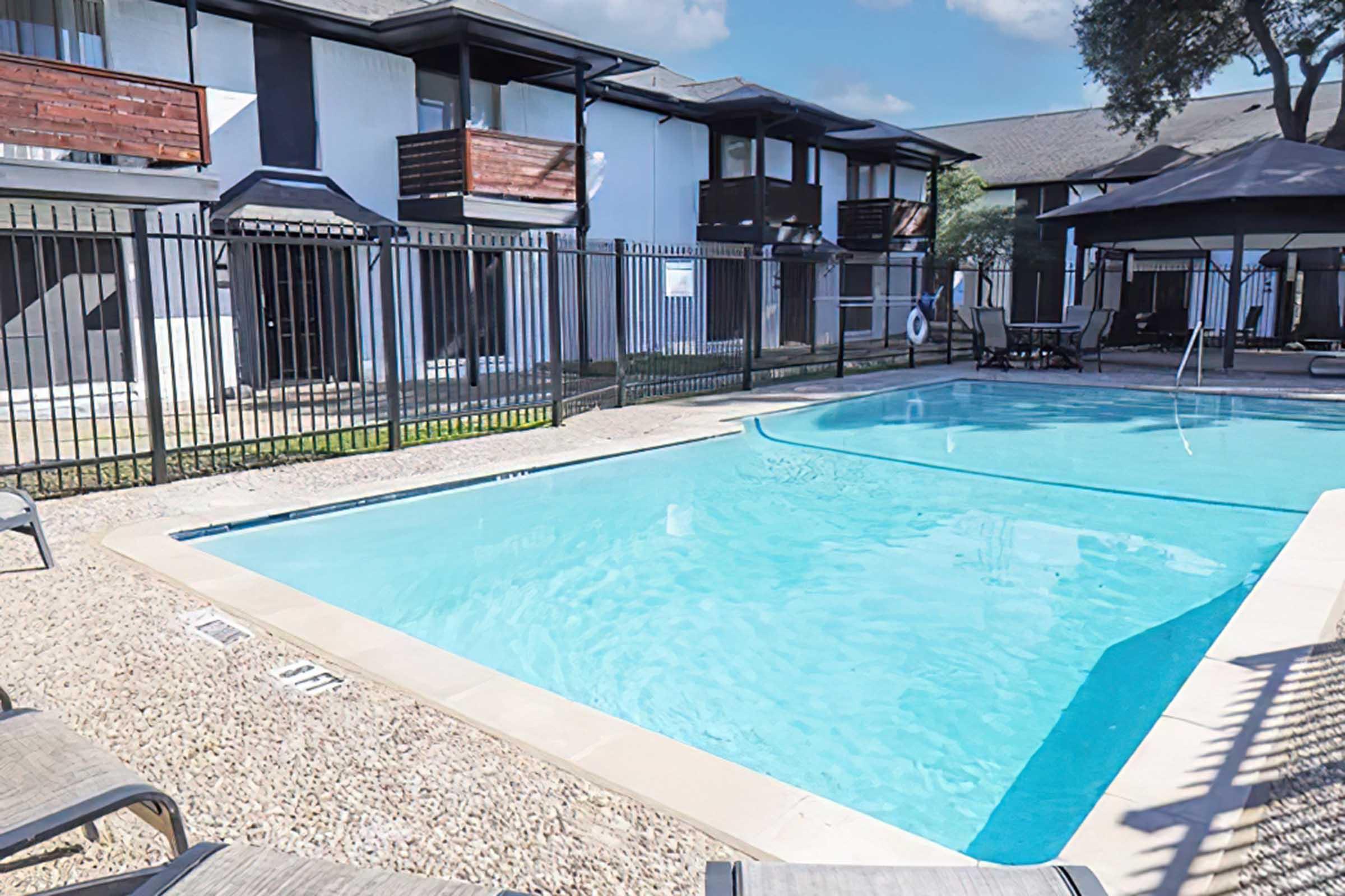 A clear swimming pool surrounded by a fenced area, with lounge chairs on the edge. In the background, there are two-story apartment buildings featuring balconies. The sky is partly cloudy, and the setting is peaceful with green grass around the pool area.