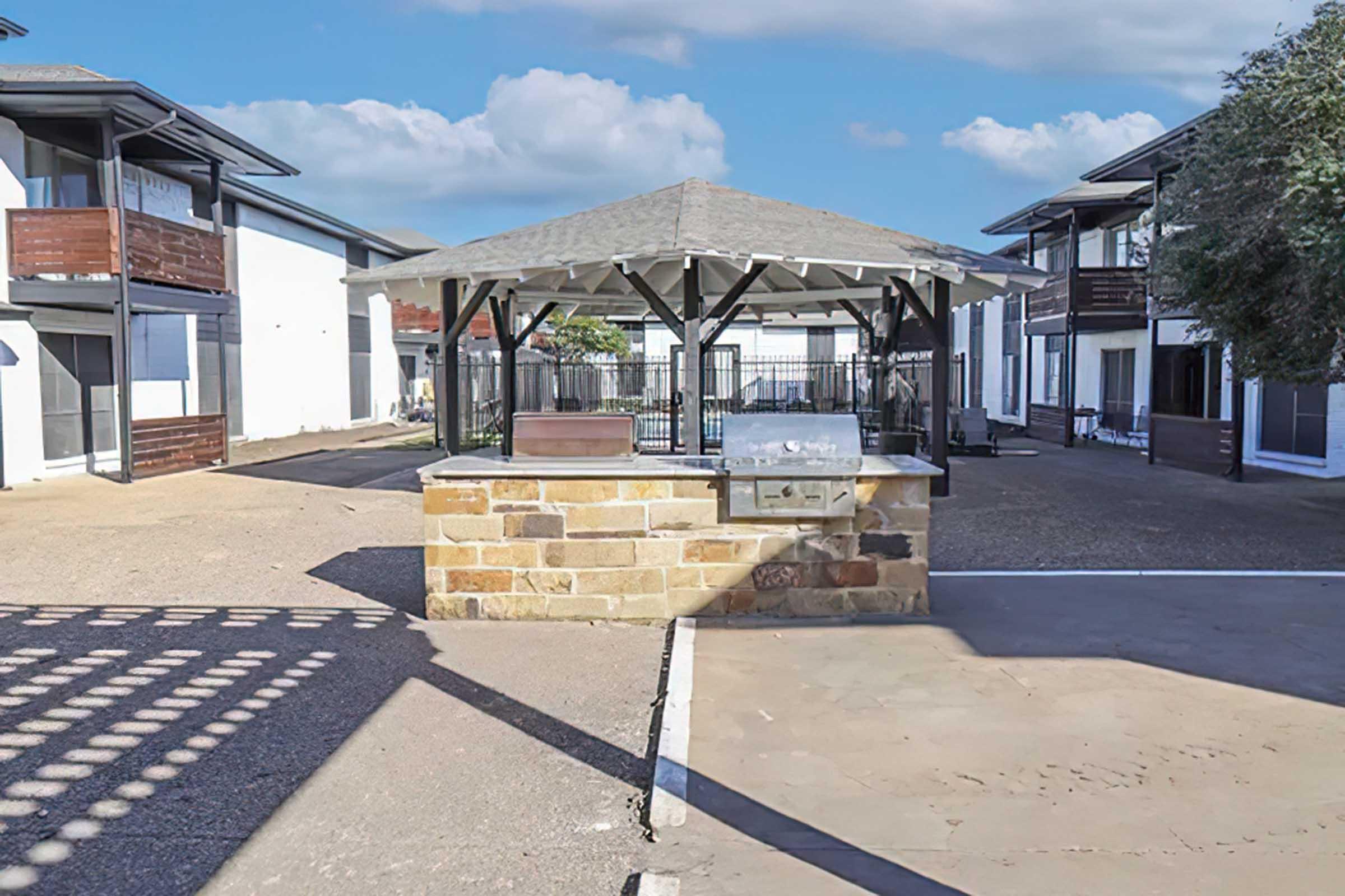 An outdoor courtyard featuring a stone barbecue grill station under a gazebo, surrounded by modern two-story buildings with balconies. The area is paved with concrete and includes a grassy patch, set against a backdrop of blue sky and scattered clouds.