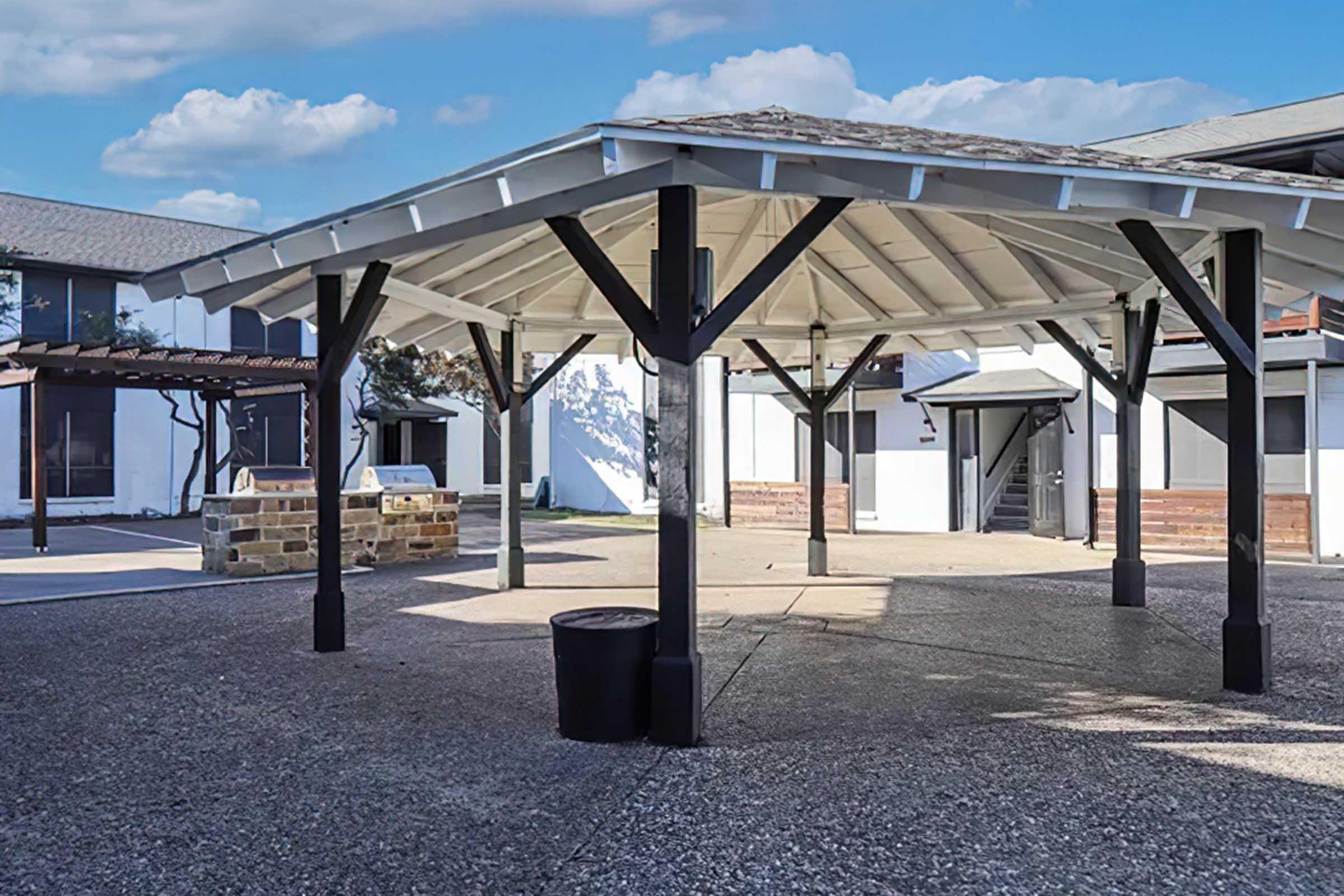 A spacious outdoor pavilion with a gabled roof supported by black wooden beams, surrounded by a courtyard area. The ground is paved, and a few buildings are visible in the background, featuring a mix of modern architecture and natural light. A trash bin is placed on the ground beneath the pavilion.