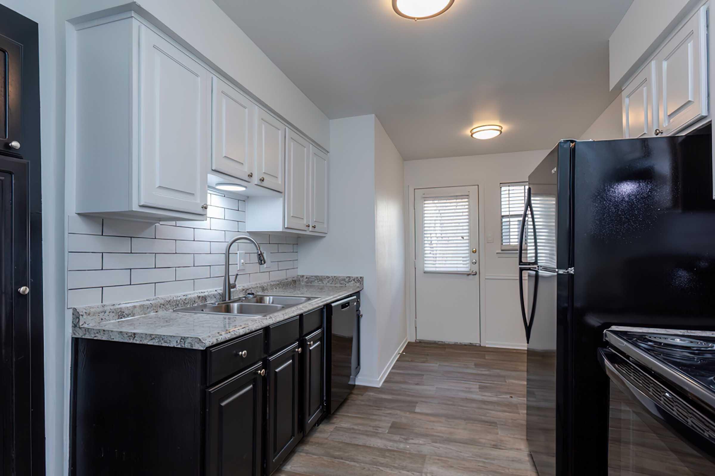 Modern kitchen with white cabinets and dark hardware, featuring a granite countertop and a stainless steel sink. Black appliances, including a refrigerator and stove, contrast with the light-colored walls and wood-like flooring. A window allows natural light, and a door leads outside.