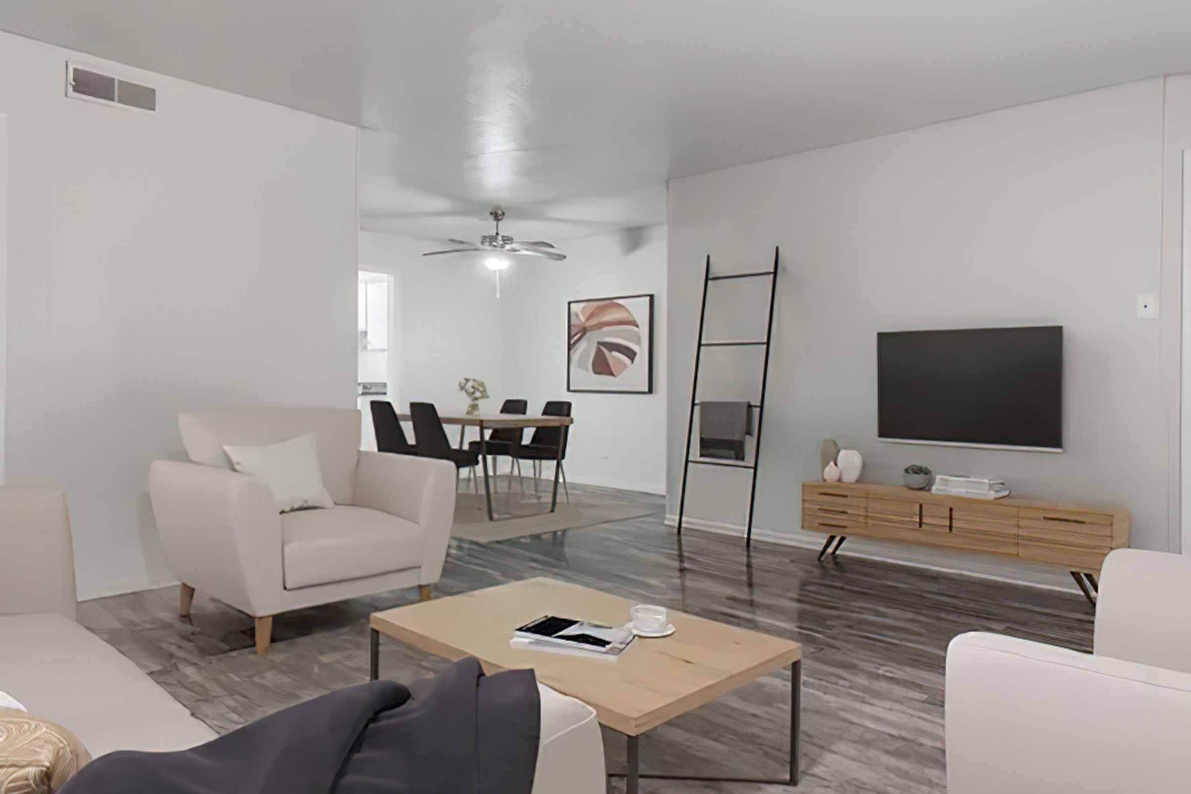 A modern living room featuring two light-colored armchairs and a coffee table. There's a wooden TV stand with a flat-screen TV and decorative items. A dining area with a table and chairs is visible in the background. The walls are light gray, and the floor has a wood-like finish.