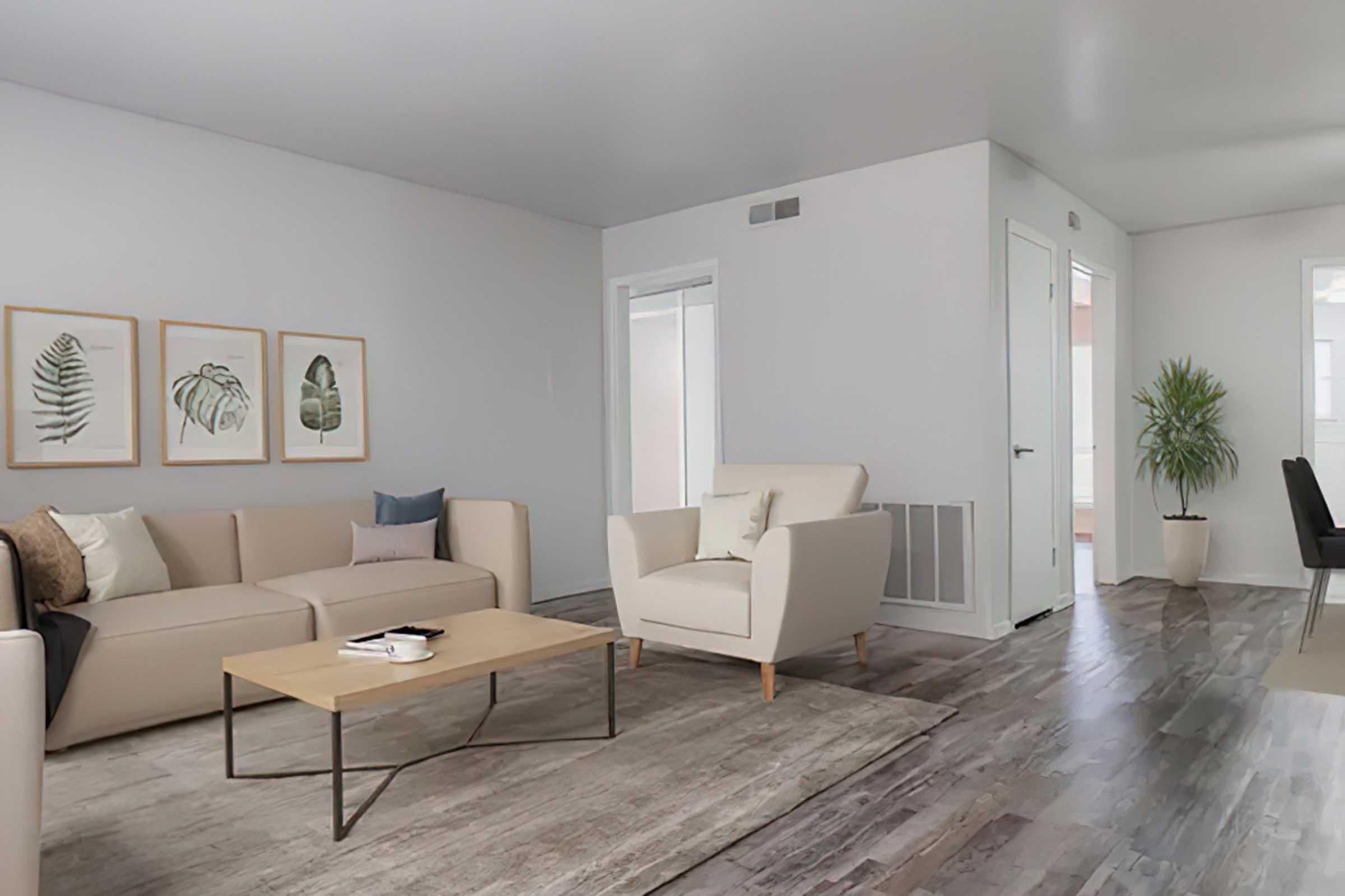 A modern living room featuring a light beige sectional sofa with decorative pillows, a small coffee table with a book, and framed botanical prints on the wall. The room has light-colored walls, a wooden floor, and a potted plant in the corner, creating a clean and inviting atmosphere.