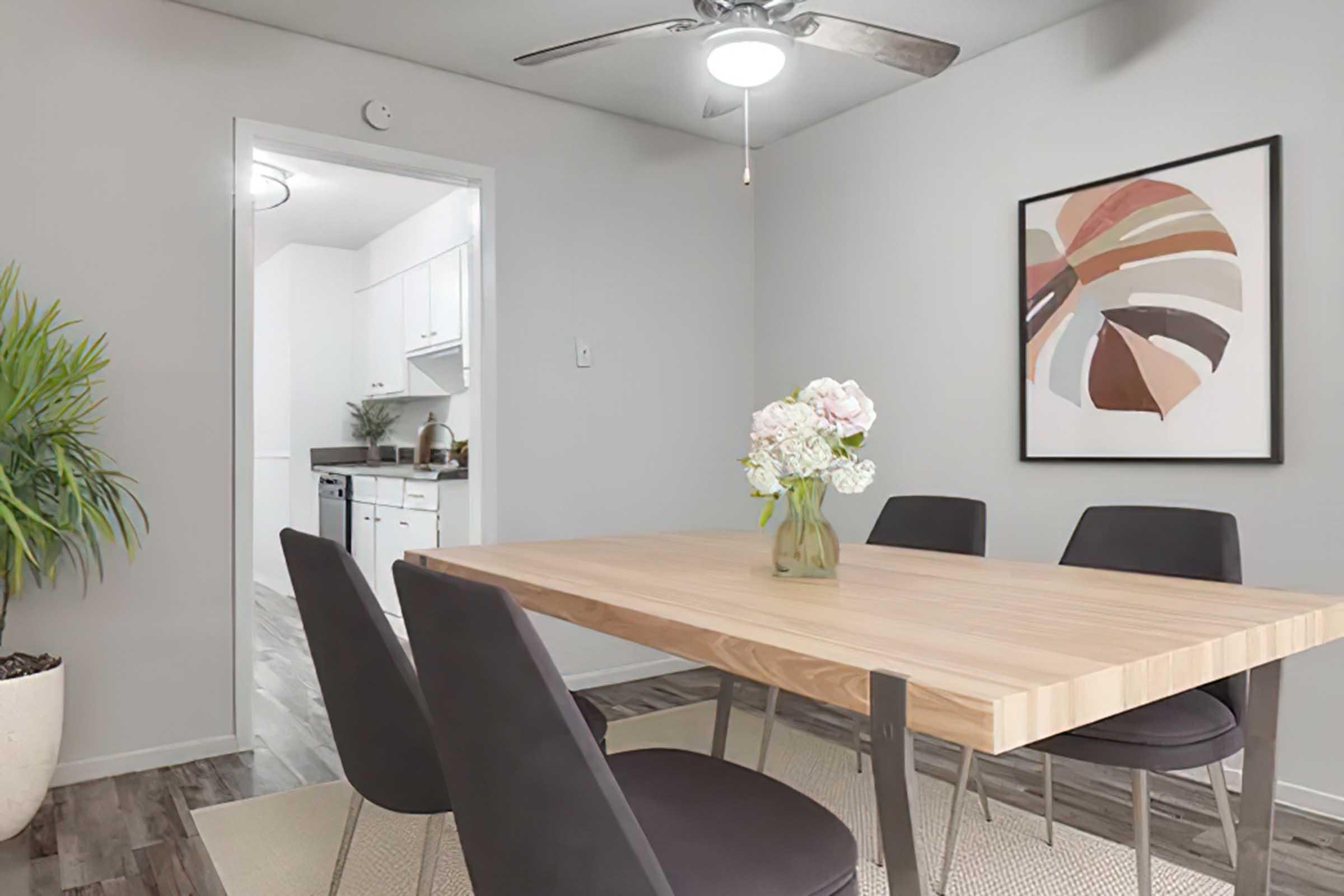 A modern dining area featuring a wooden table with four black chairs. A vase with flowers sits on the table. A large wall art piece is displayed, showcasing abstract shapes in muted colors. A doorway leads to a kitchen, and a potted plant is placed in the corner, adding a touch of greenery.