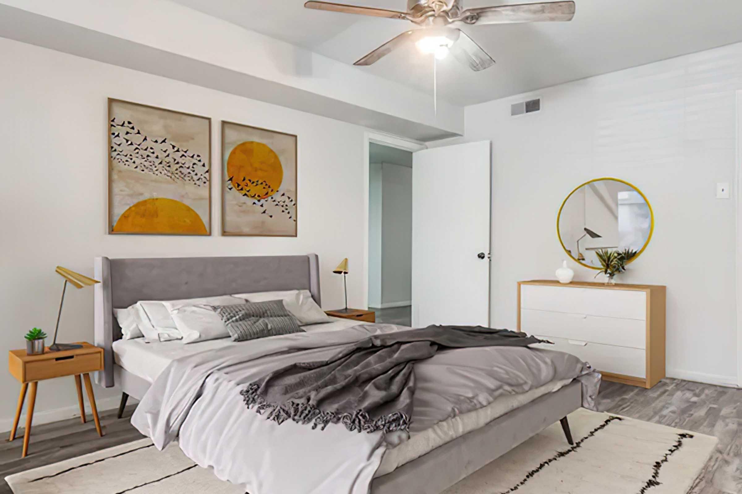 A modern, cozy bedroom featuring a gray bed with a textured throw blanket, decorative pillows, and a nightstand with a lamp. On the wall hang two framed prints of birds and suns. A round mirror is mounted above a dresser, with minimal decor items. Soft lighting from a ceiling fan creates a warm ambiance.