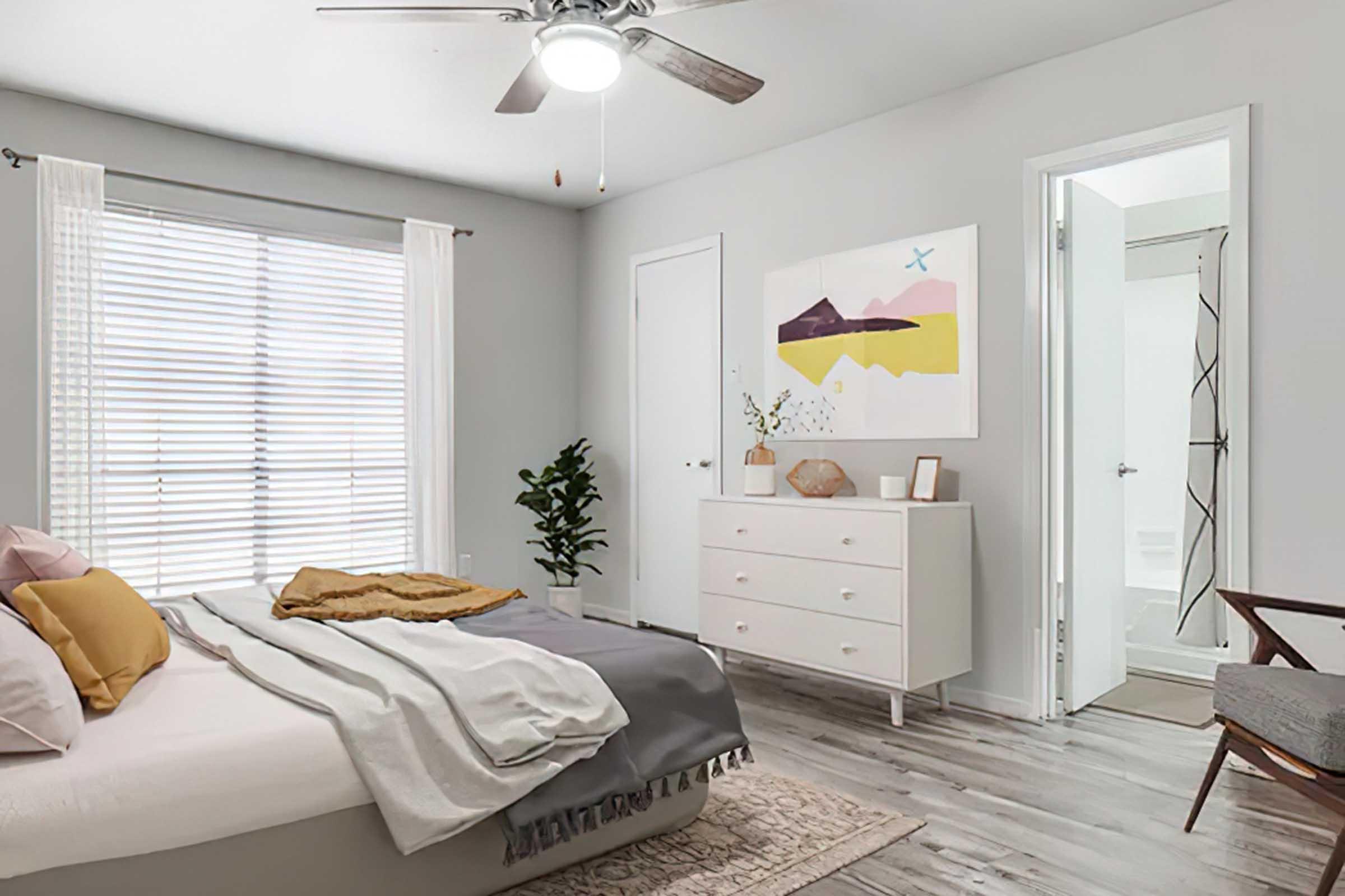 A modern bedroom with a neatly made bed featuring purple and yellow pillows. A decorative throw is draped over the foot of the bed. There is a white dresser against the wall and a potted plant nearby. The room has light gray walls, a ceiling fan, and a large window with white blinds. A bathroom door is visible in the background.