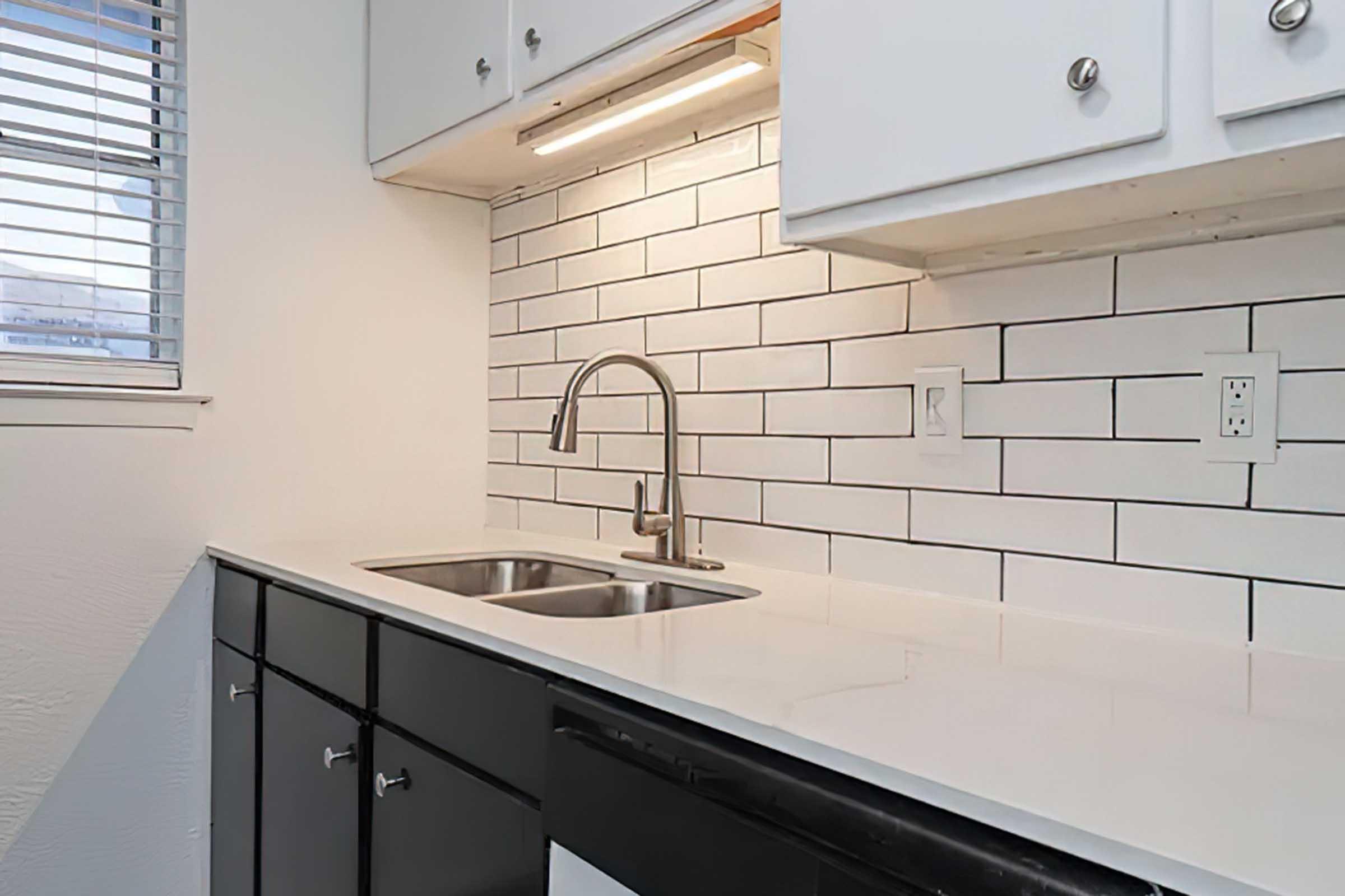 Modern kitchen with a white tiled backsplash, stainless steel faucet above a double sink, and dark cabinetry. A light fixture is installed under the cabinets, illuminating the countertop. The window features blinds that allow natural light to enter.