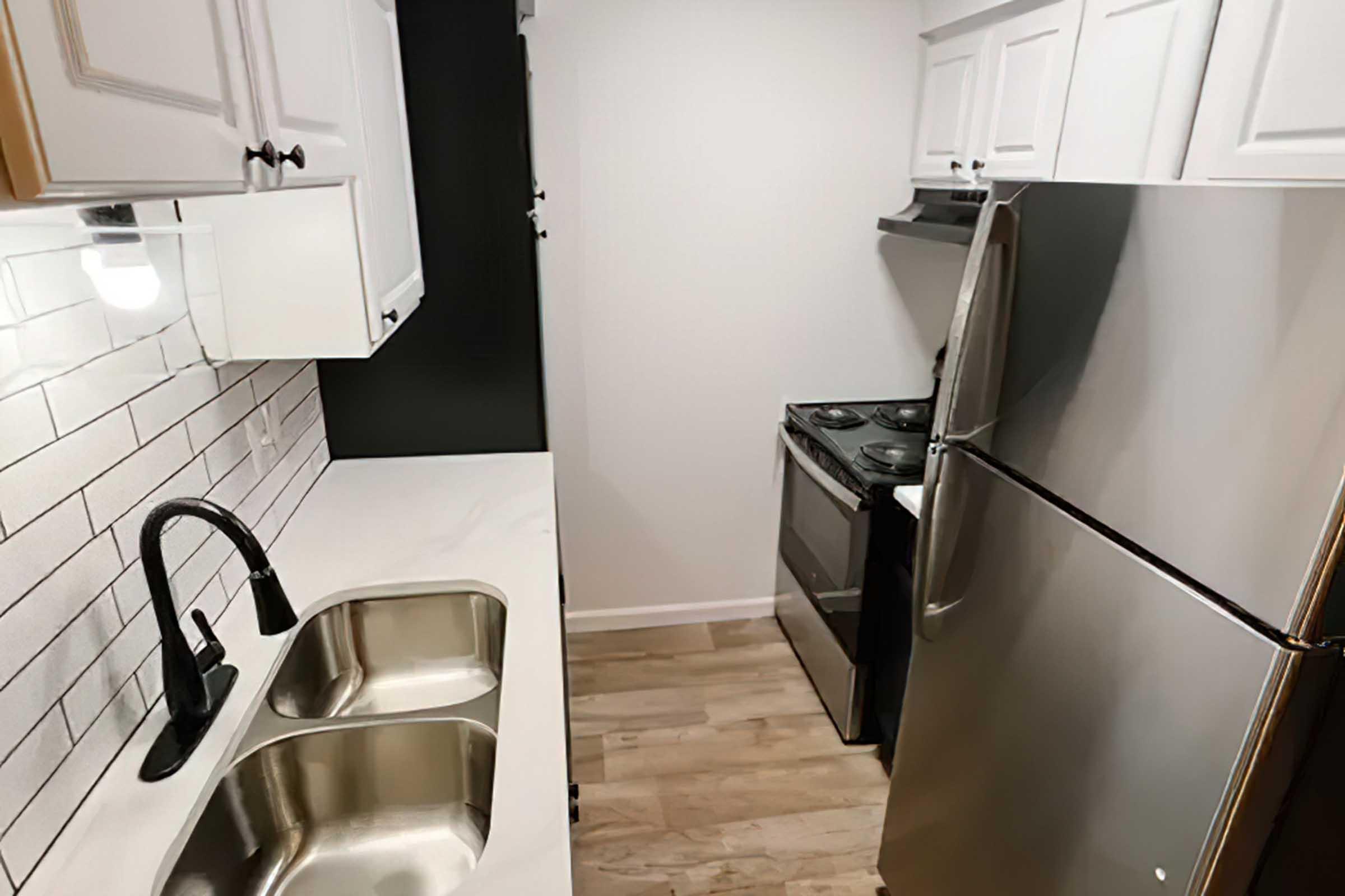 A modern kitchen featuring white cabinets, a stainless steel refrigerator, and an oven. The countertops are white with a minimalist design, and there's a double sink with a sleek black faucet. The walls have a patterned backsplash with gray tiles, and the flooring is light wood.