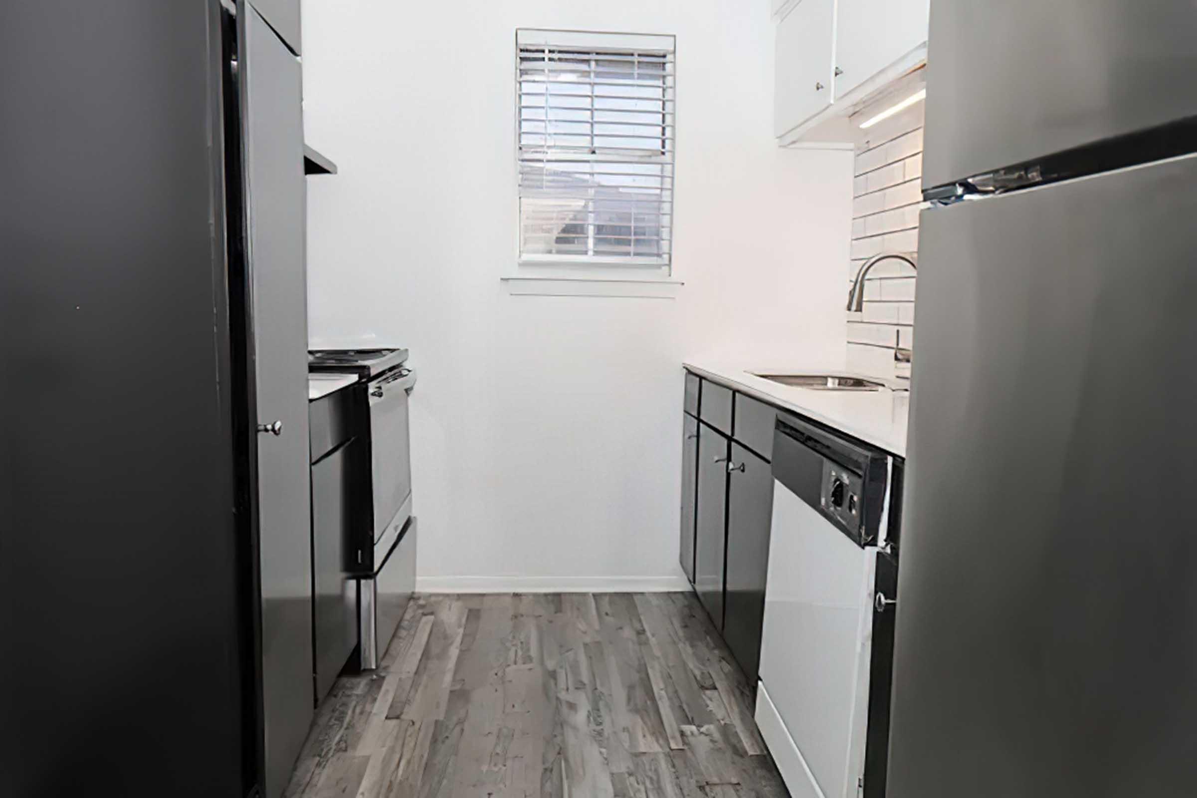 A modern kitchen with black appliances, including a stove and refrigerator. A white countertop complements dark cabinetry. There's a window with blinds allowing natural light, and the floor is finished with light wood-like tiles. The design is sleek and contemporary.