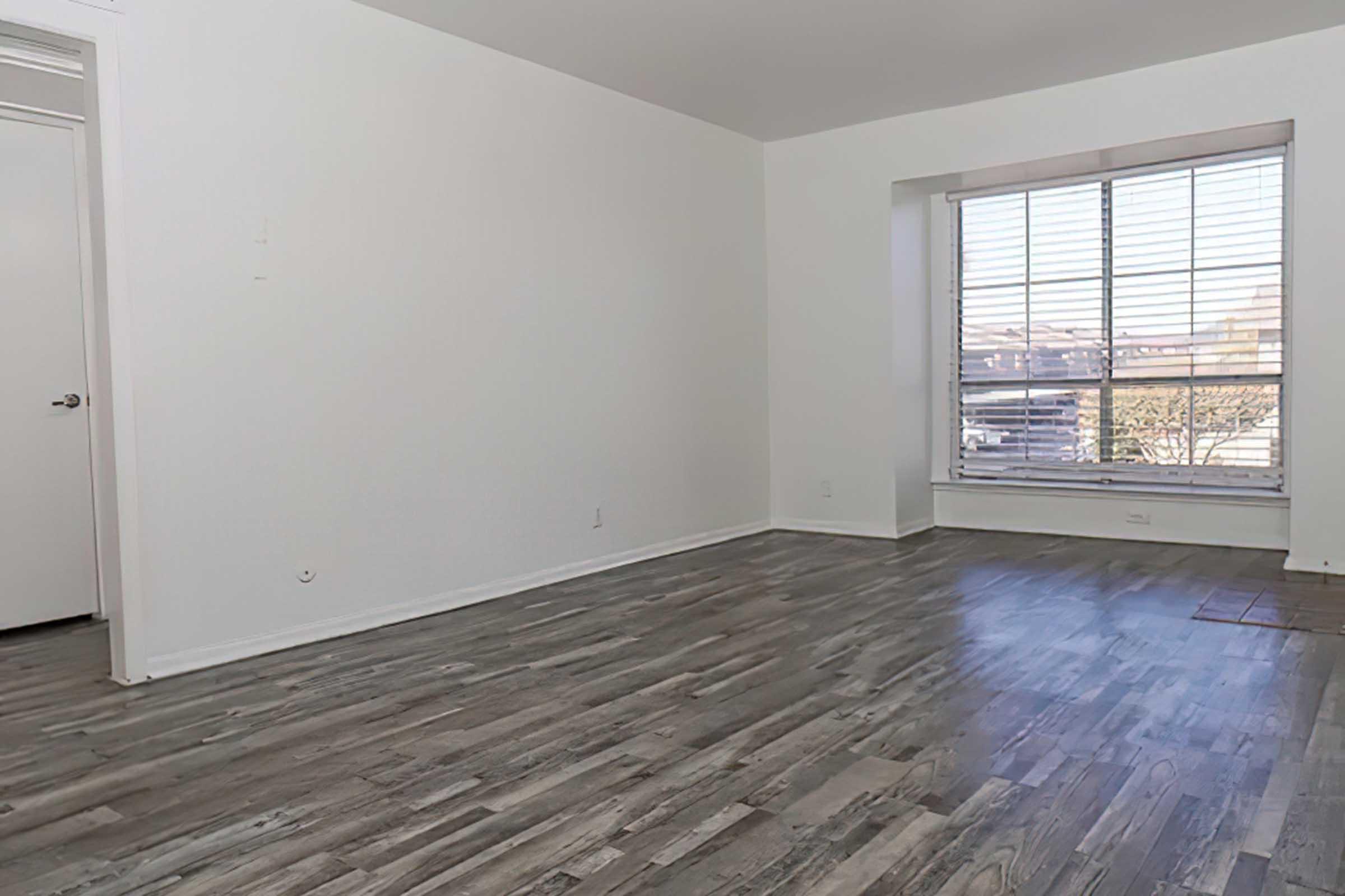 A spacious, empty room featuring light-colored walls and a large window with blinds. The floor is covered in light wooden laminate. There is a door on the left leading to another space, and sunlight is streaming in through the window, brightening the area.
