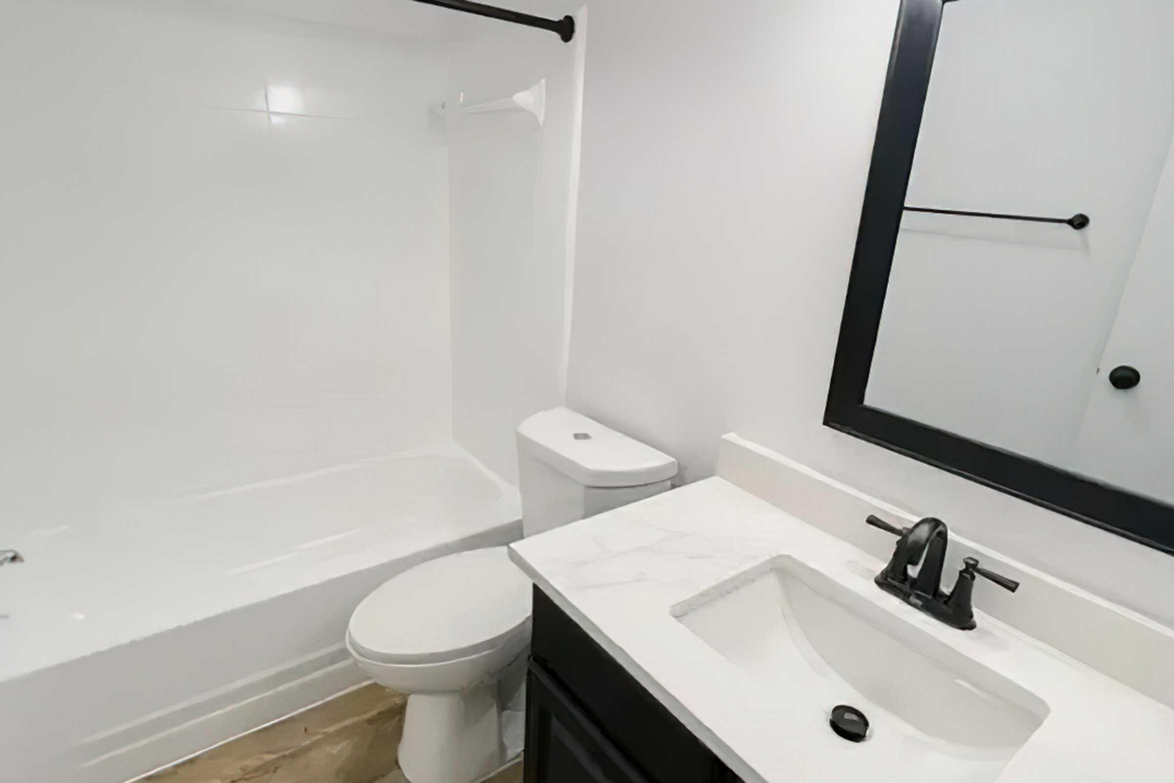 A modern bathroom featuring a white tub, toilet, and a sleek black-framed mirror above a marble countertop with a sink. The walls are painted white, and the floors have a neutral tile design, creating a clean and contemporary aesthetic.