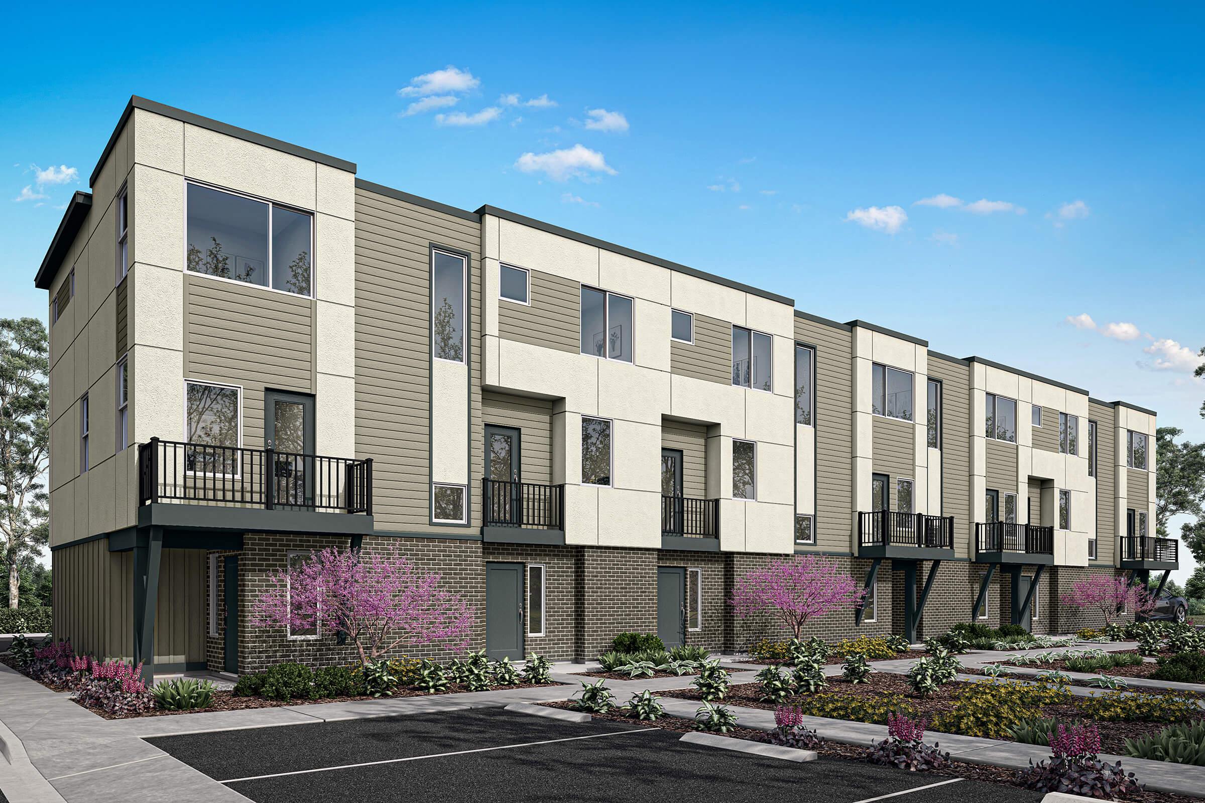 A modern row of townhouses featuring a mix of light-colored siding and brick exteriors. Each unit has large windows and private balconies. The architecture is contemporary, surrounded by landscaped gardens with blooming flowers, under a clear blue sky. Parking spaces are visible in front of the townhouses.
