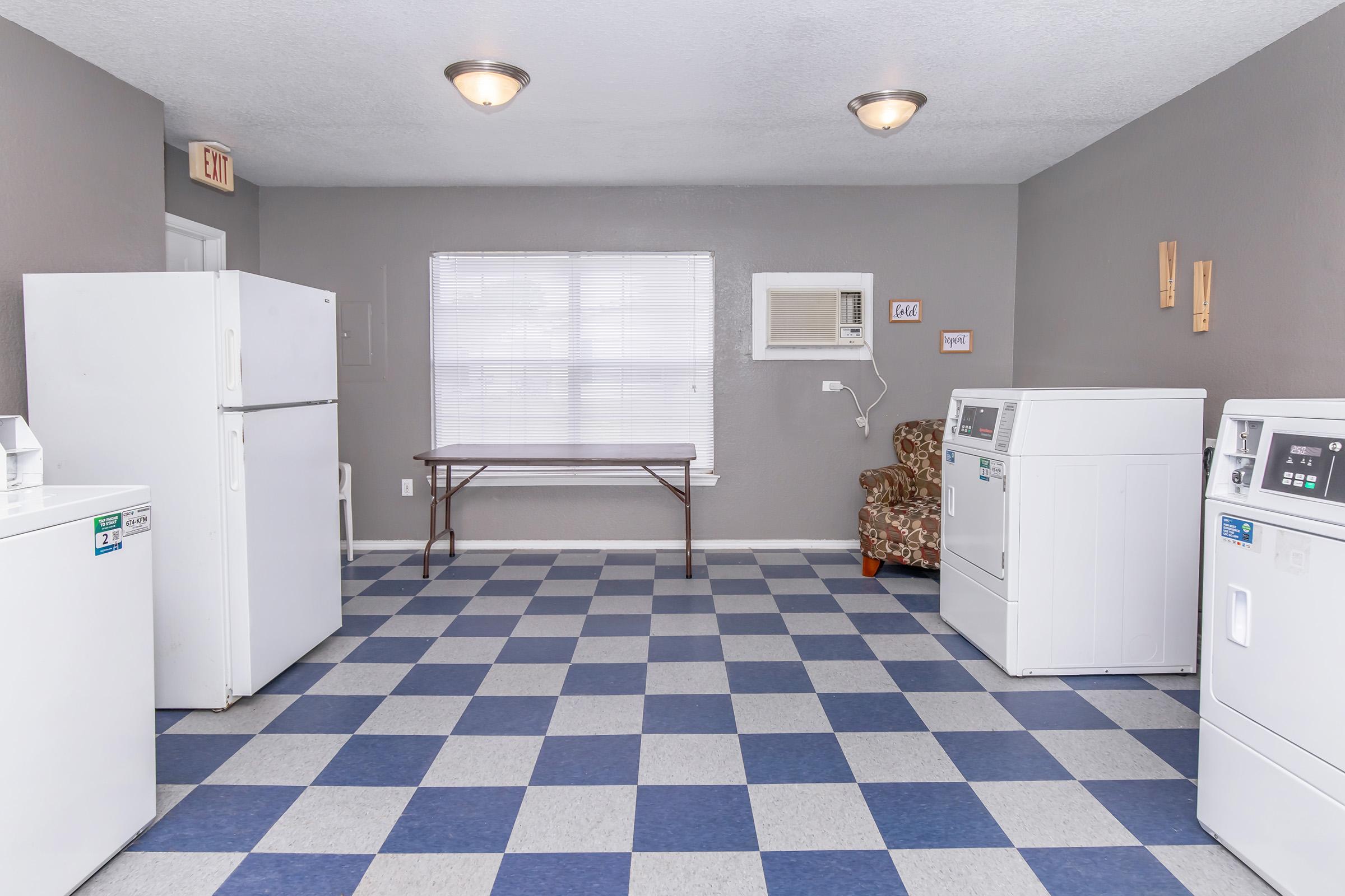 A clean, well-lit laundry room featuring a white refrigerator, a table against the wall, and two washing machines. The floor has a blue and white checkered pattern, while a comfortable chair is visible in the corner. Windows provide natural light, enhancing the spacious feel of the room.