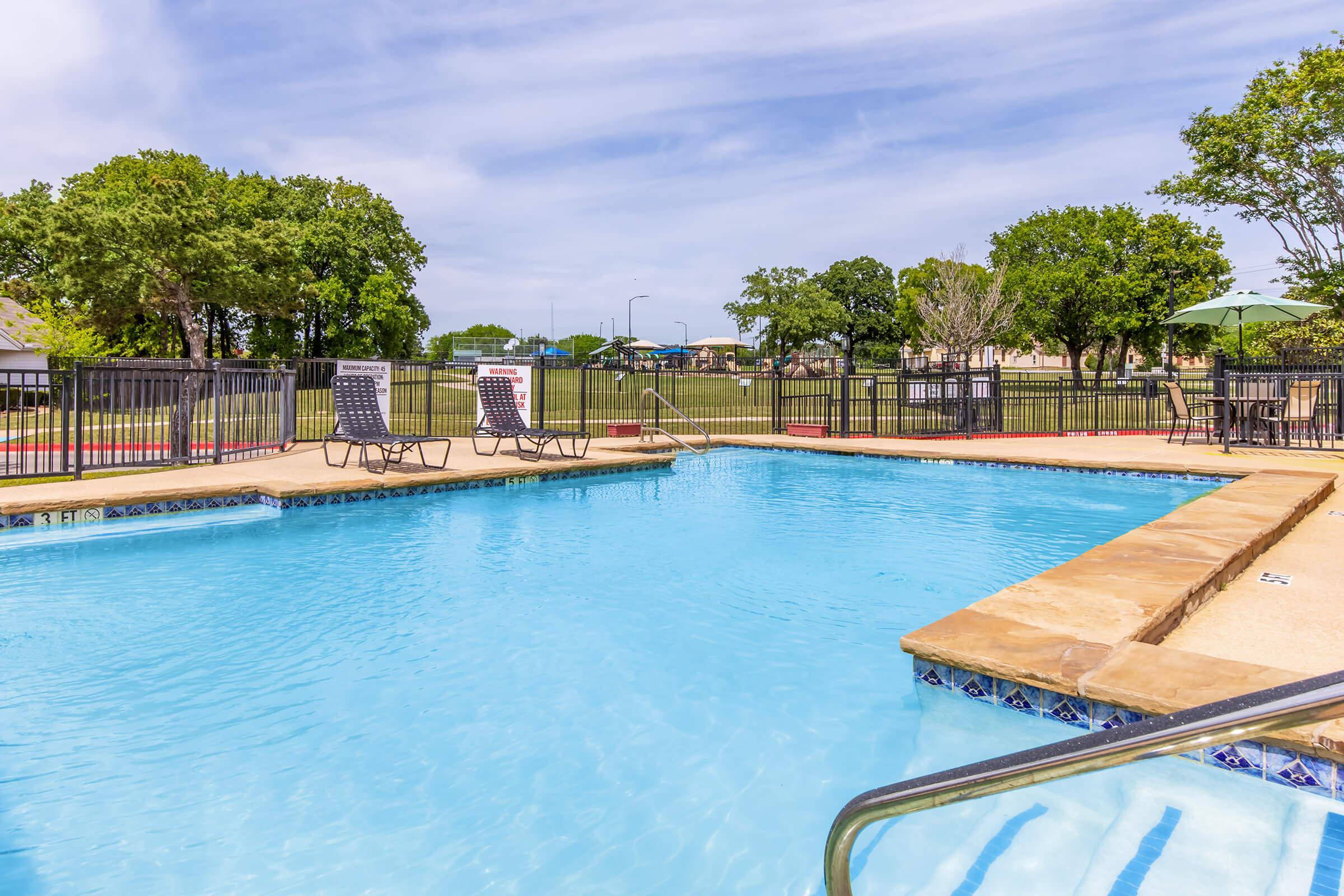 A clear blue swimming pool surrounded by a tan deck and black metal fencing. There are several lounge chairs nearby and green trees in the background. A grassy area is visible beyond the pool, creating a relaxing outdoor environment.