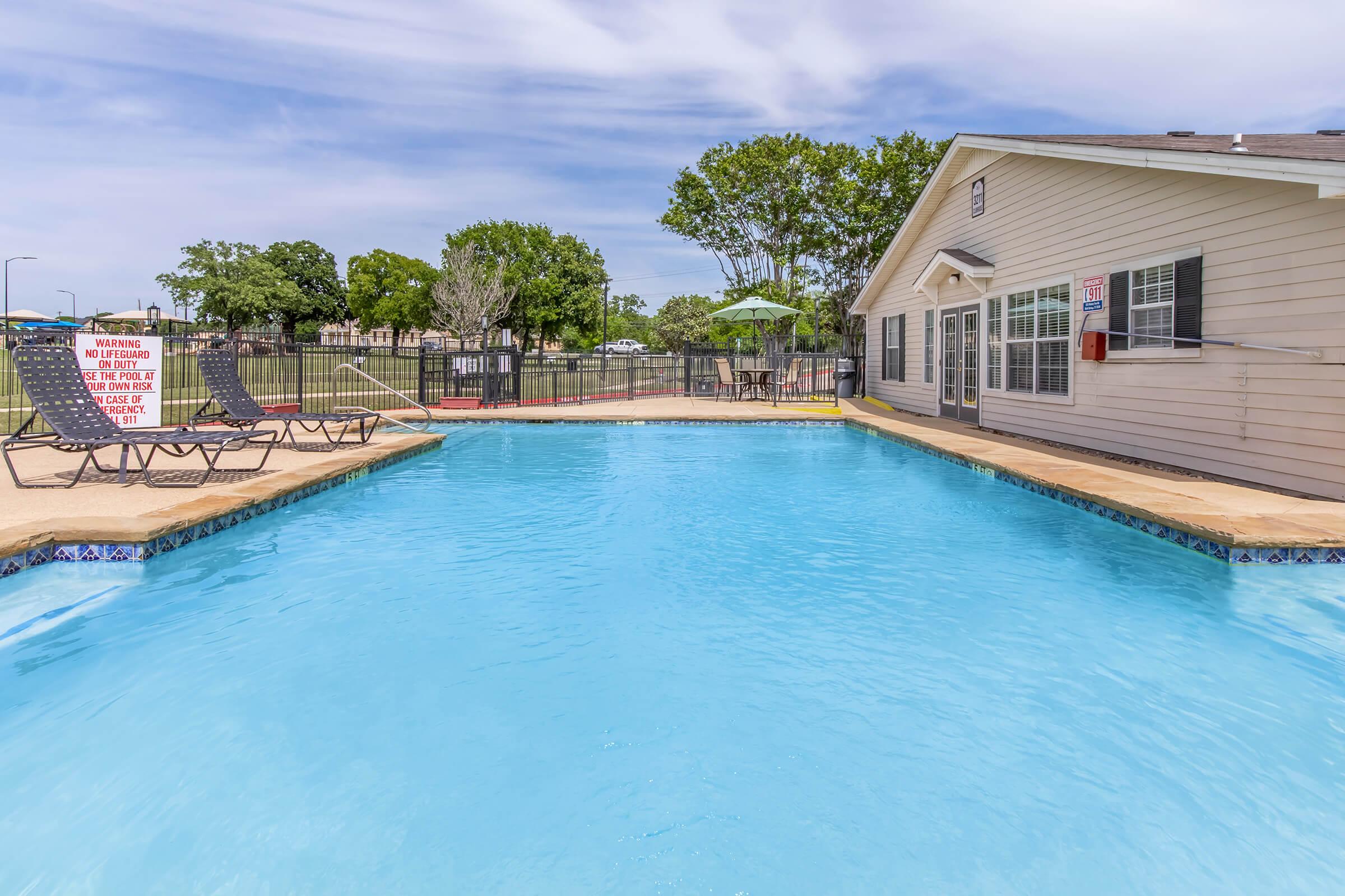 A clear blue swimming pool surrounded by lounge chairs and an adjacent building. In the background, there are trees and a fenced area. A sign warns of pool rules. The setting is bright and inviting, suggesting a recreational atmosphere.