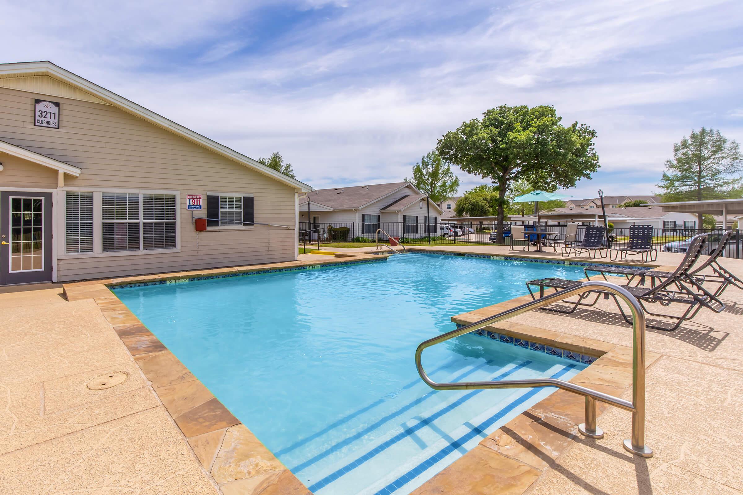 A clear blue swimming pool with a gently sloping entry, surrounded by lounge chairs and tables. In the background, there is a single-story building with several windows and a door, along with trees and greenery. The sky is partly cloudy, contributing to a bright, inviting atmosphere.