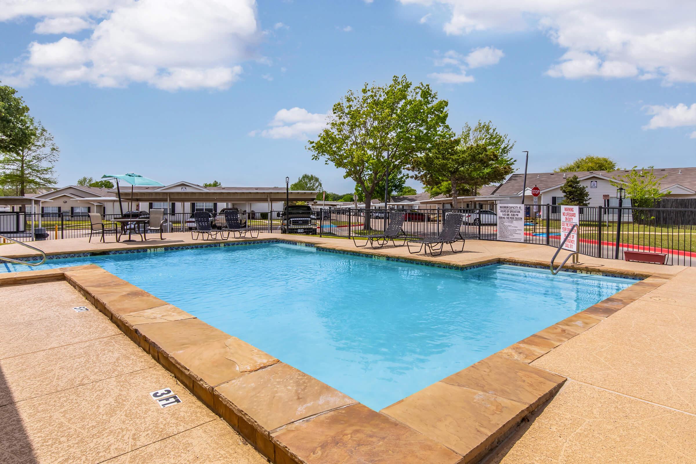 A clear swimming pool with a light blue surface, surrounded by a concrete deck and lounge chairs. Trees are visible in the background, along with houses and a cloudy blue sky. Poolside signs warning about depth and safety are also present.