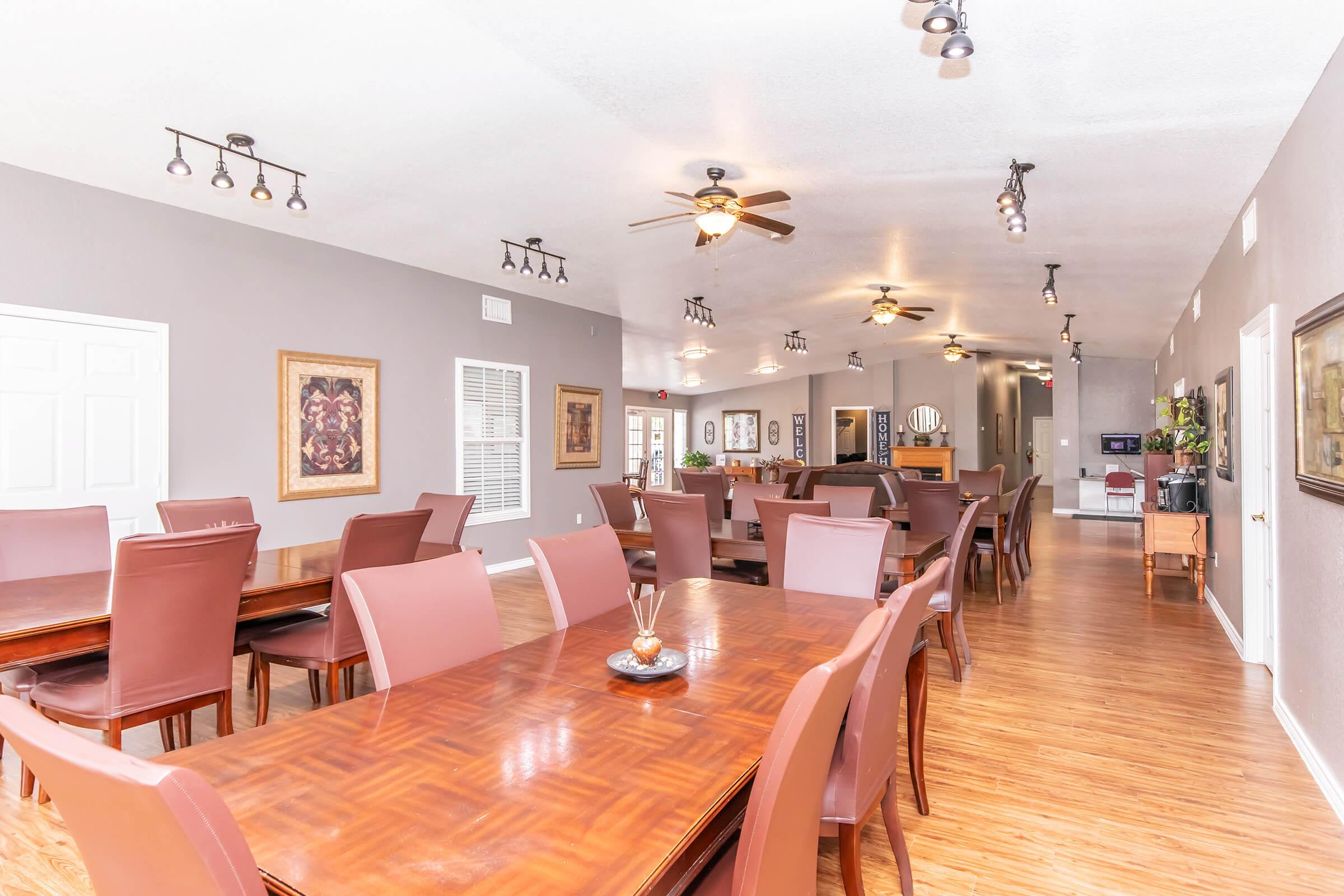 A spacious dining area with multiple wooden tables and chairs, featuring soft lighting from ceiling fixtures. The walls are painted in neutral tones and adorned with artwork. The room is designed for gatherings, showcasing an inviting atmosphere with a combination of modern and classic decor.