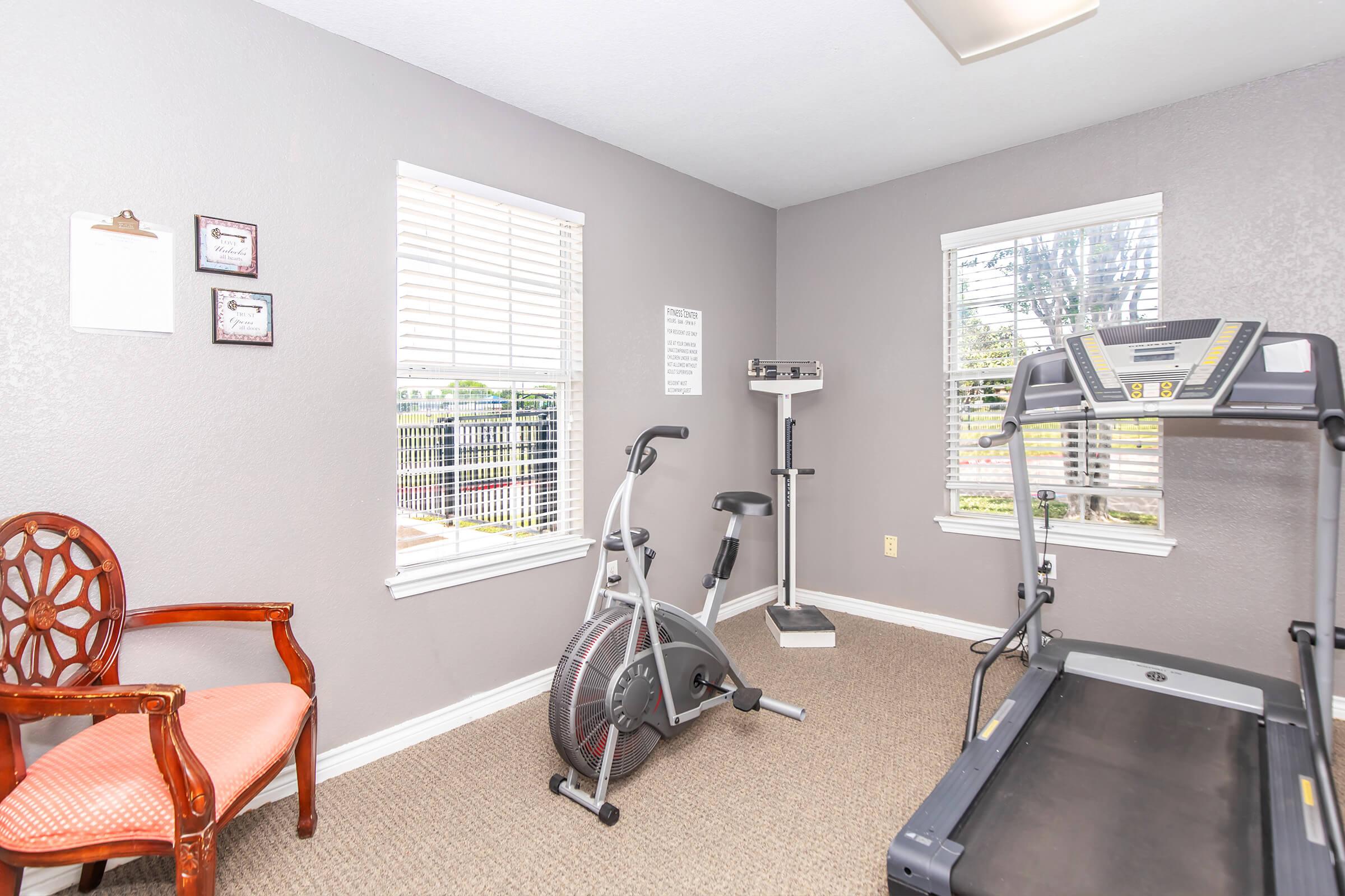 Home gym with a treadmill, exercise bike, and a scale. There’s a chair with an orange cushion and framed pictures on the wall. Two windows allow natural light, and the room has gray walls and carpeted flooring.