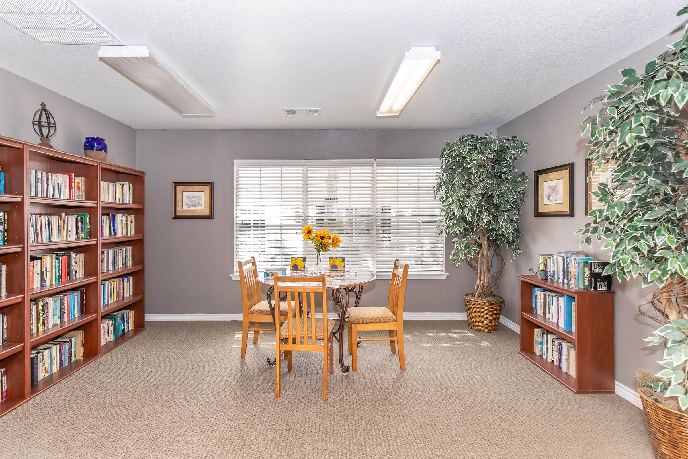 A cozy reading nook featuring two wooden chairs and a small round table surrounded by bookshelves filled with books. Sunflower centerpieces adorn the table, and large windows allow natural light to illuminate the space. Lush potted plants add greenery to the room, creating a warm, inviting atmosphere.