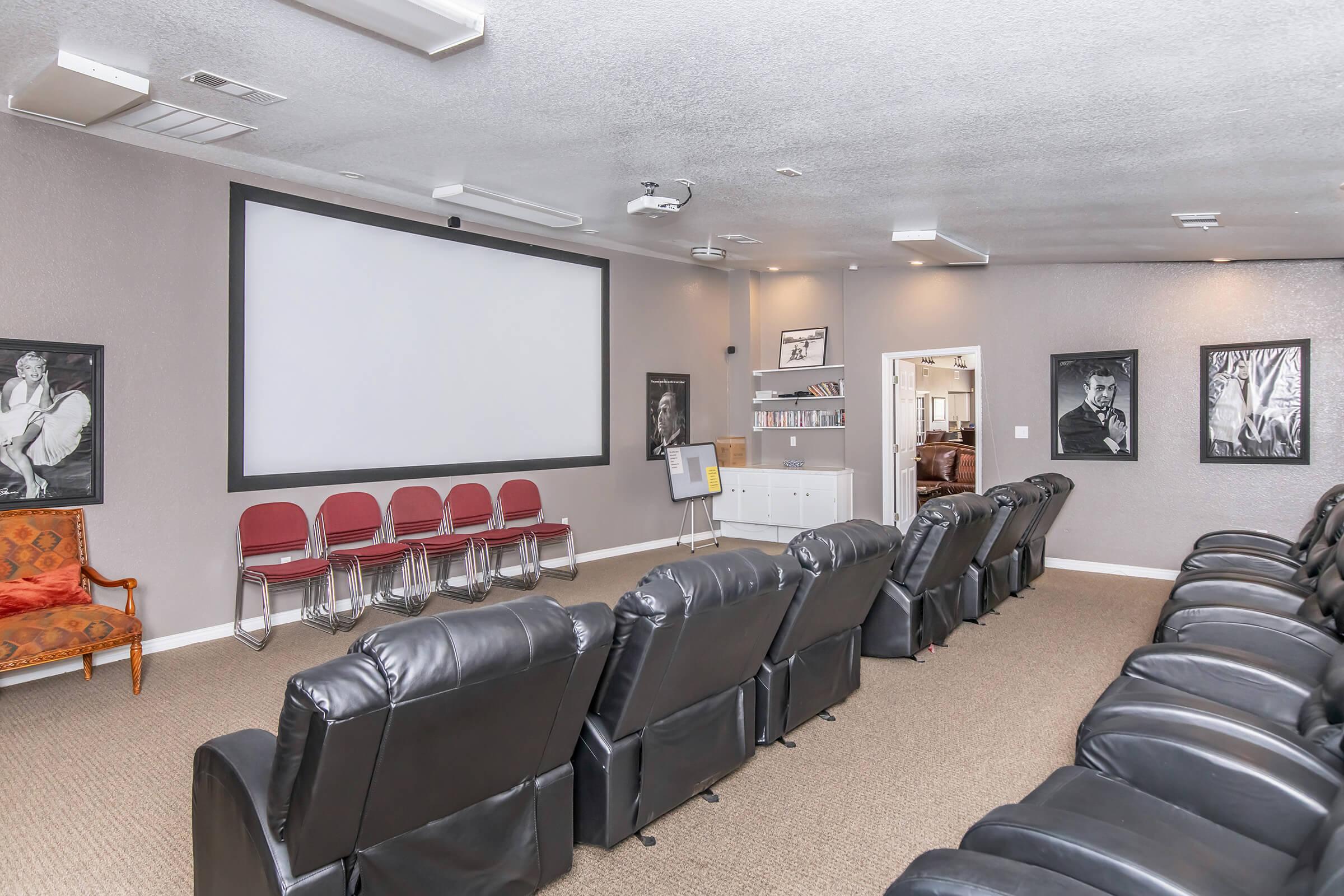 A home theater room featuring dark leather recliner seating arranged in rows, a large projection screen at the front, and framed black-and-white portraits on the walls. There are several empty red chairs and a small bookshelf in the background, creating a cozy and inviting movie-watching environment.