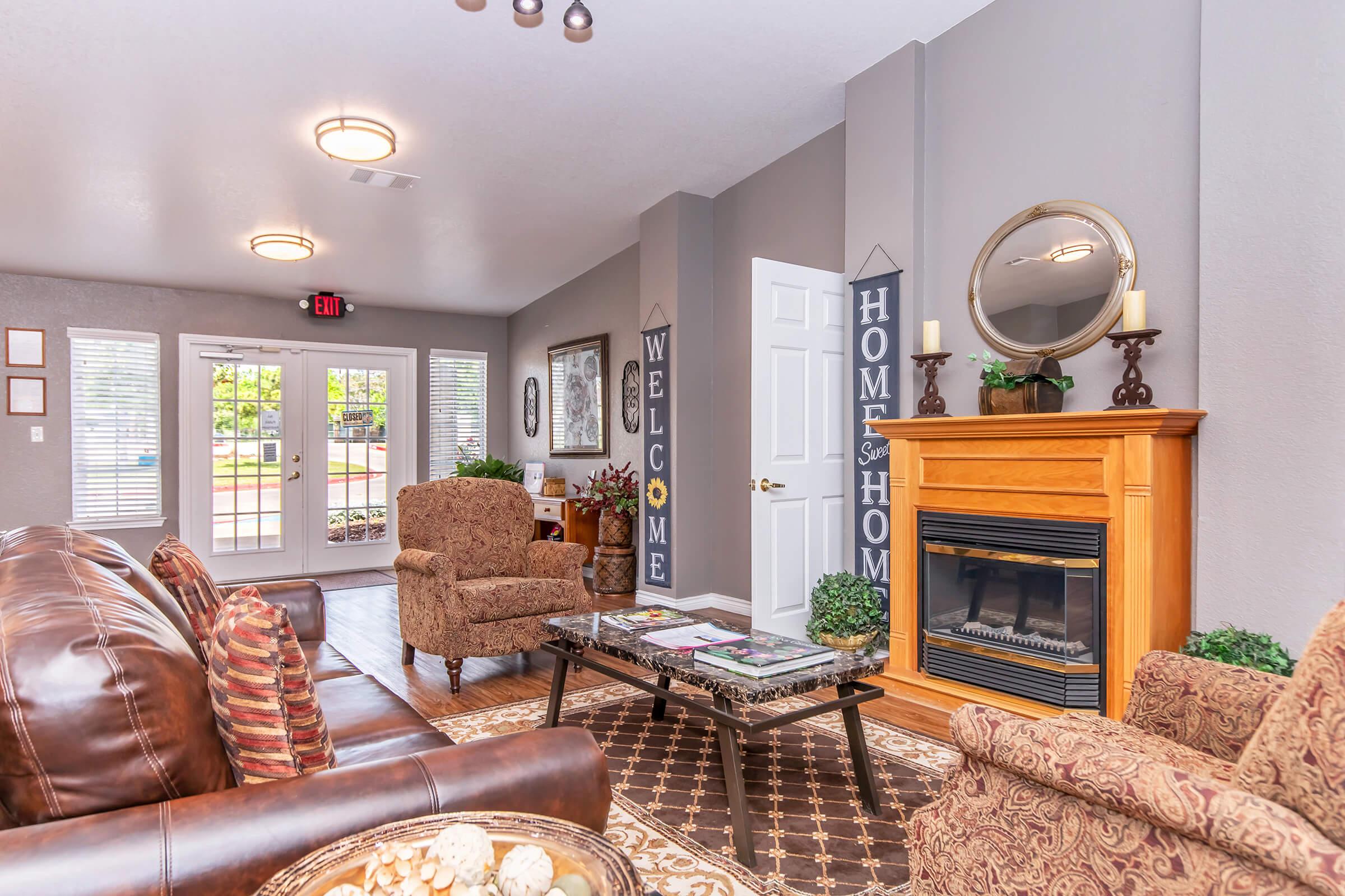 A cozy living room featuring brown leather sofas, a coffee table, and a wooden fireplace. Decorative plants and a mirror adorn the space. Walls are painted in soft gray, with "Welcome" and "Home Sweet Home" signs. Large windows allow natural light, and the entrance leads outside.