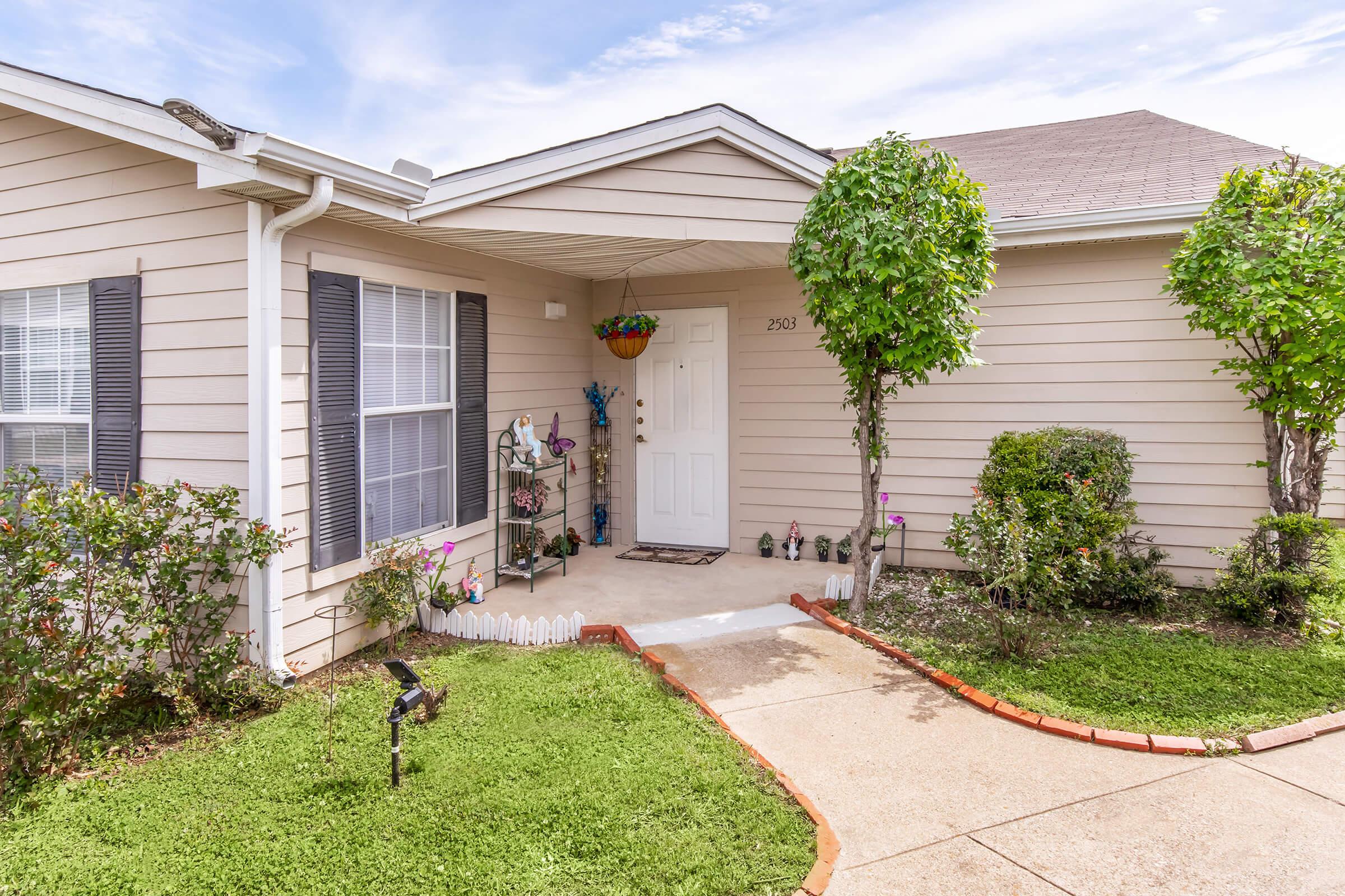 A small, well-kept house with light-colored siding and a welcoming front porch. The yard features neatly trimmed grass, a few decorative plants, and a potted flower arrangement. There's a white picket fence section and stone pathway leading to the front door, creating an inviting atmosphere.