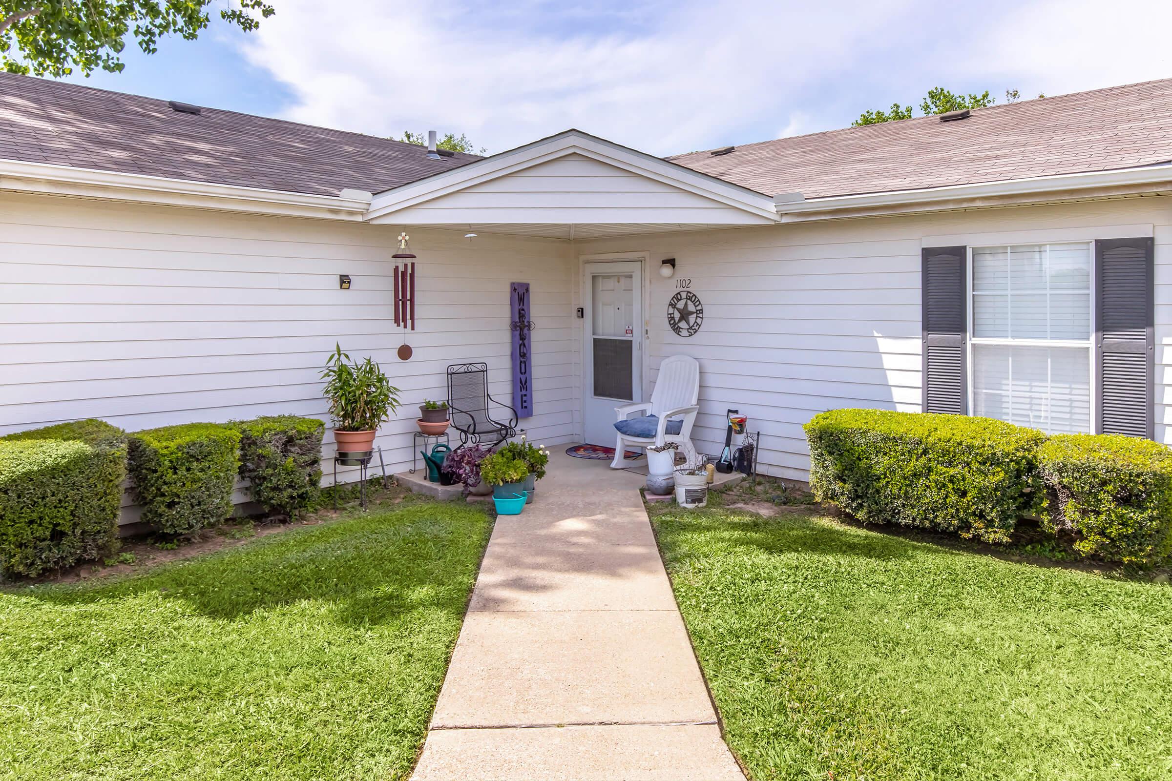 A welcoming front porch featuring a white house with gray shutters. A path leads to the door, surrounded by neatly trimmed hedges and potted plants. Decorative elements include a welcome sign and a wind chime hanging near the entrance, with a chair placed on the porch for relaxation.