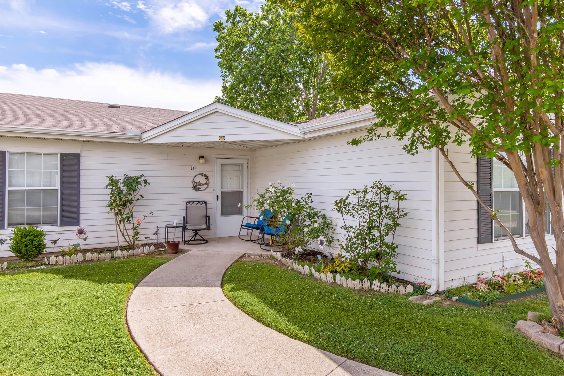 A charming front yard featuring a well-maintained lawn, bordered by a white picket fence. The house has a light-colored exterior with gray shutters and a welcoming porch with seating. Lush green bushes and flowering plants enhance the inviting atmosphere under a bright blue sky.