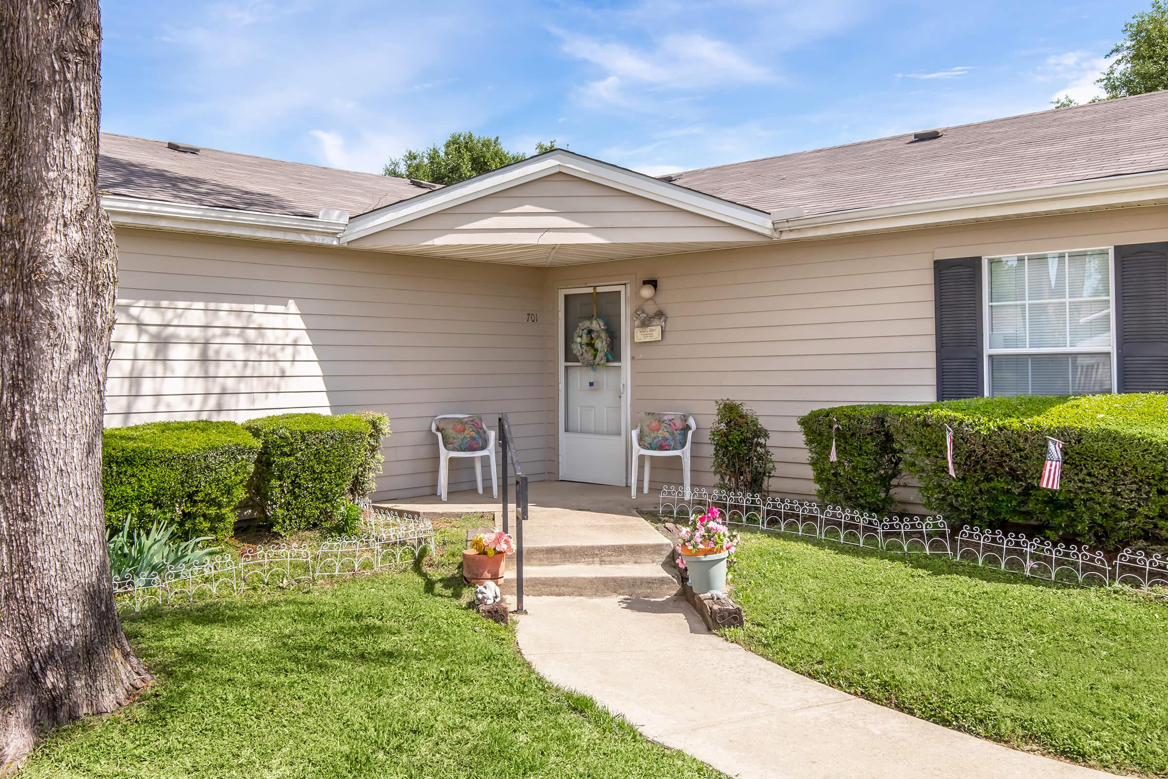 A single-story house with a light-colored exterior, featuring a front door with decorative elements and two white chairs on the porch. The well-maintained yard has a green lawn, hedges, and flower pots, creating a welcoming atmosphere. A clear blue sky is visible in the background.