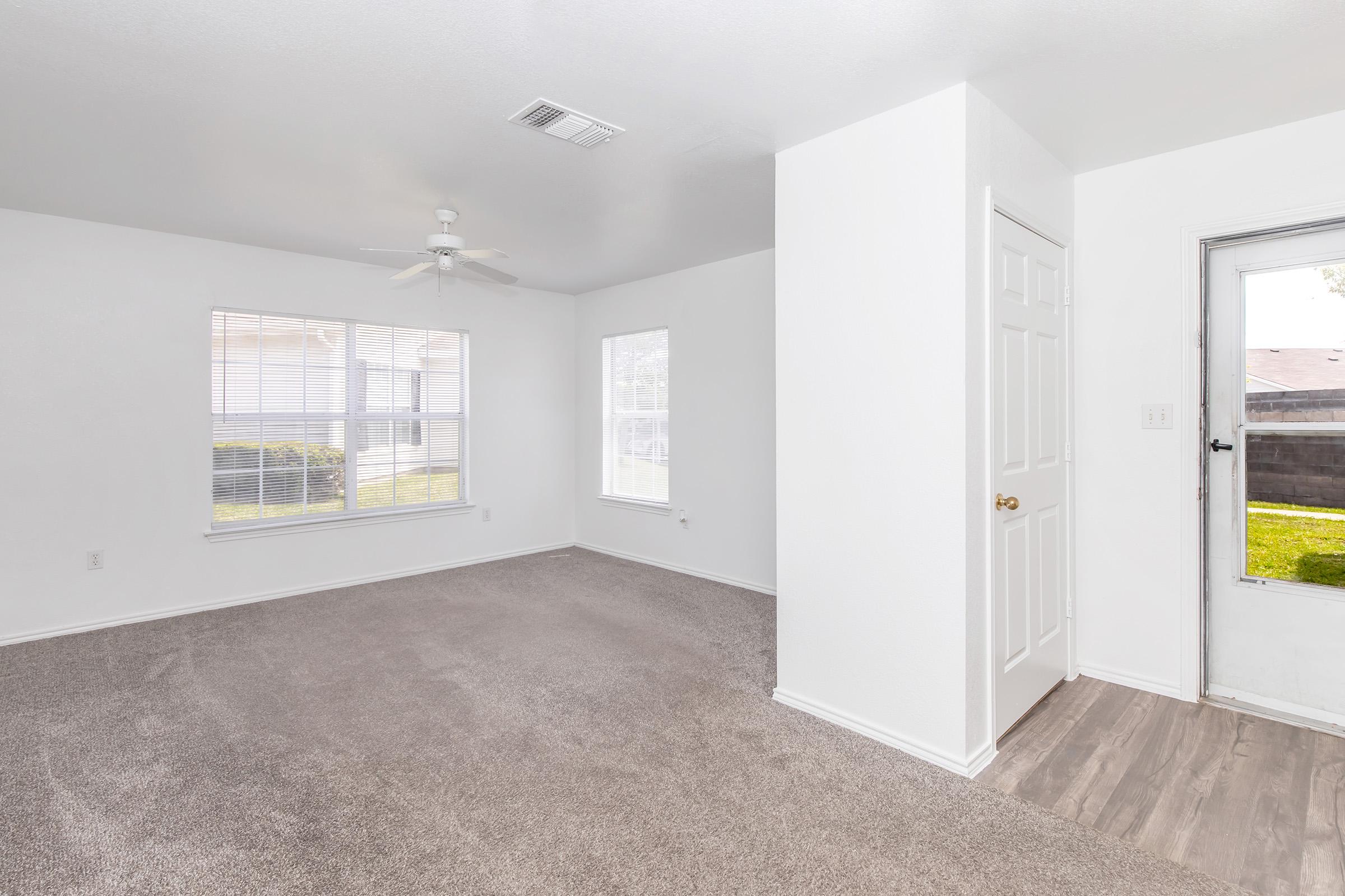 A bright, inviting living room with light-colored walls and carpet. It features a ceiling fan, two large windows allowing natural light, and a door leading outside. The space is clean and uncluttered, suitable for various layouts and designs.