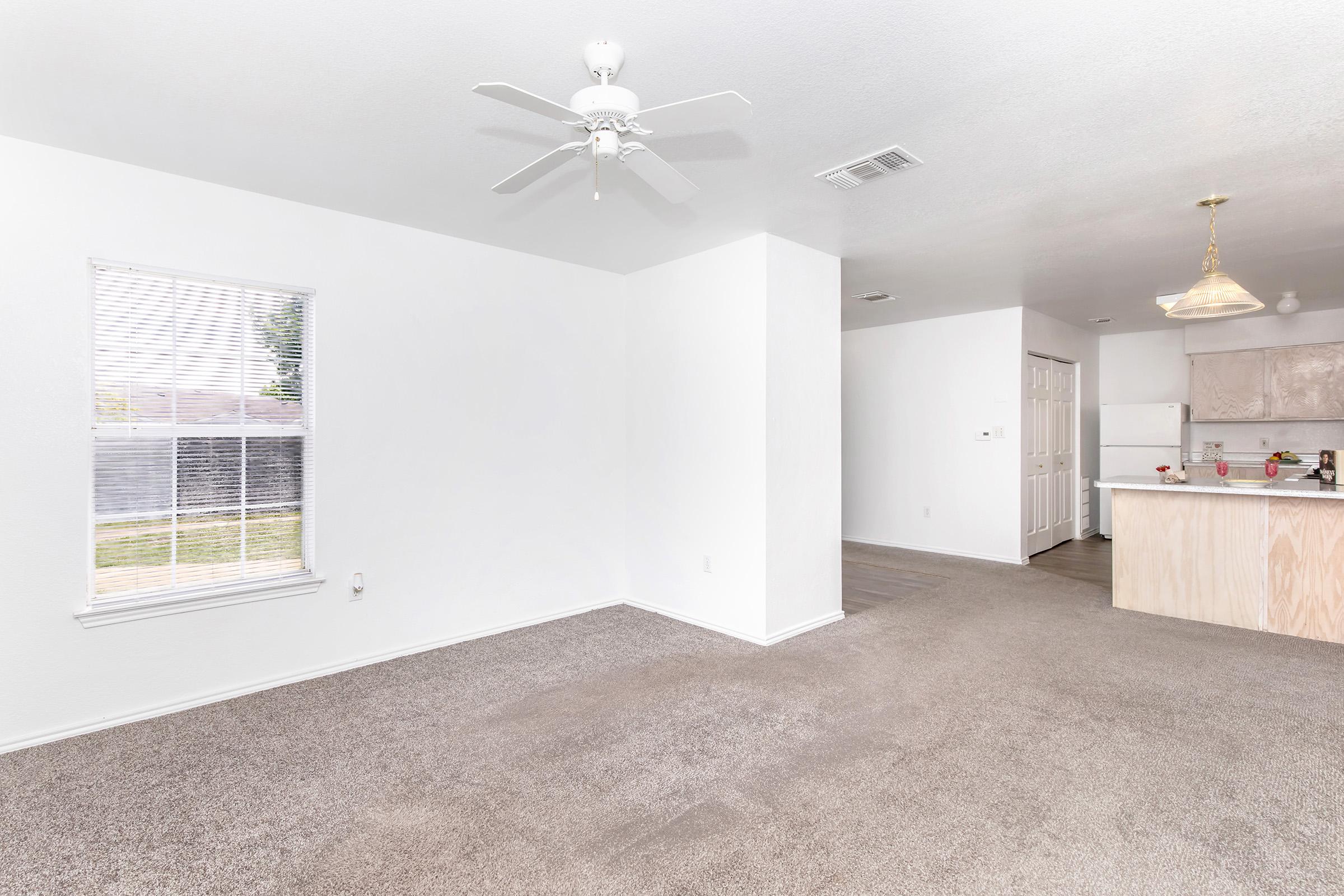 A bright, spacious living room with light brown carpet and white walls. There's a ceiling fan and a large window allowing natural light. An adjacent kitchen with light wooden cabinets is partially visible in the background, featuring an open layout that connects the two spaces.