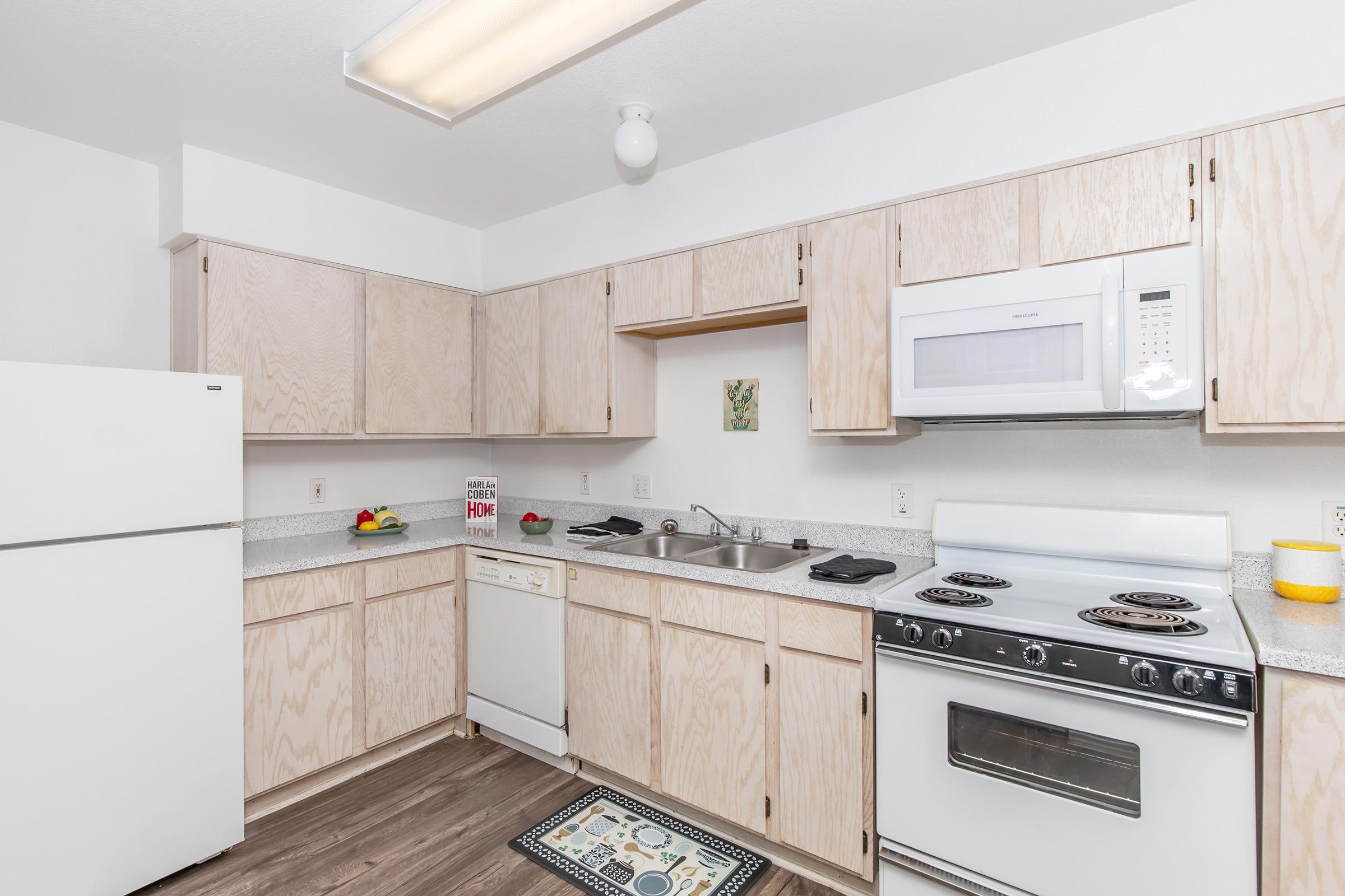 A modern kitchen featuring light wood cabinetry, a white refrigerator, a dishwasher, and a white microwave above a gas stove. The countertop is light gray with a small rug on the floor. There are some decorative items and kitchen utensils visible, creating a clean and functional space.