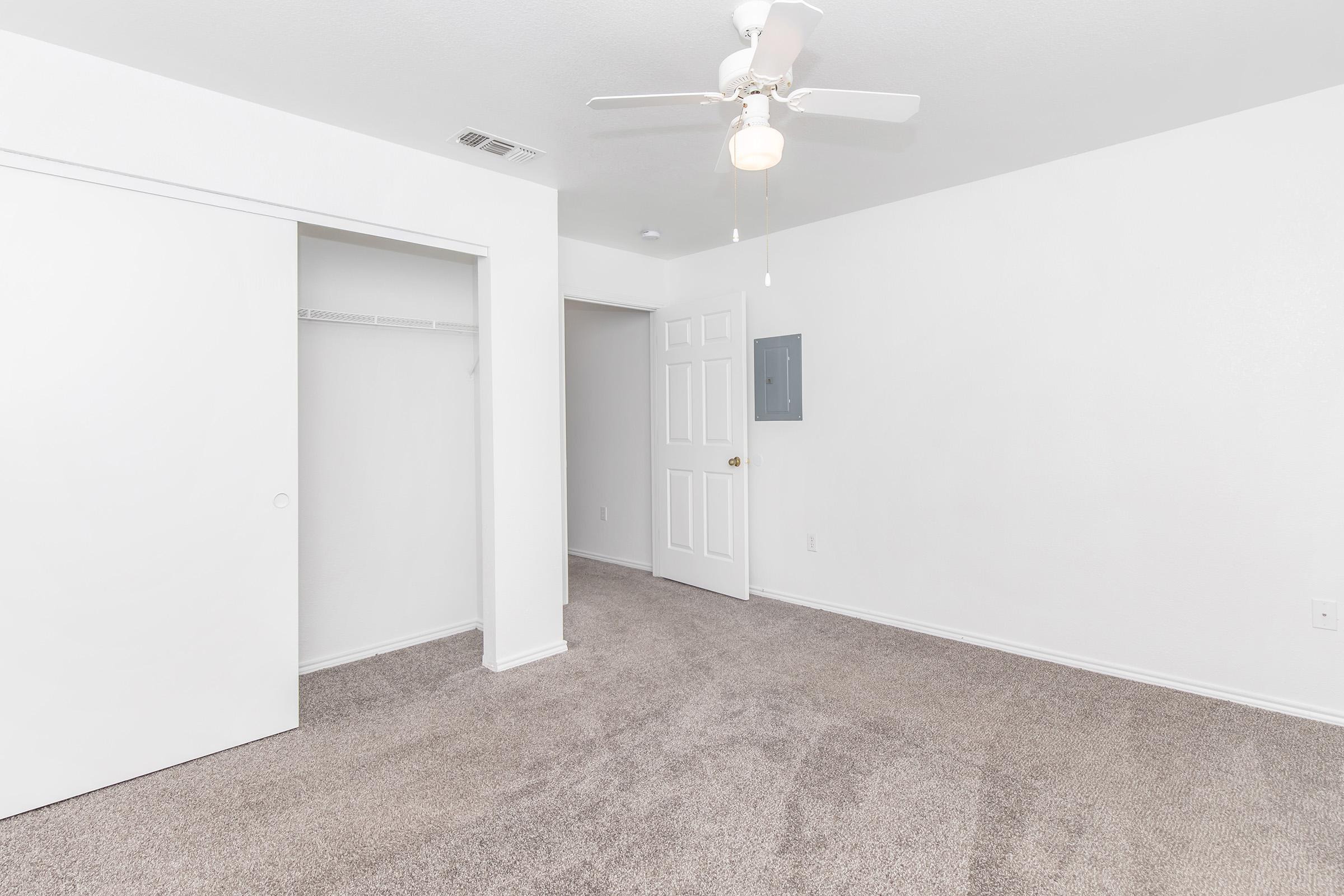 A clean, empty bedroom featuring light-colored walls and carpet. There is a ceiling fan with a light fixture, a white closet with sliding doors, and a door leading to another room. The space is well-lit and invites a sense of openness and simplicity.