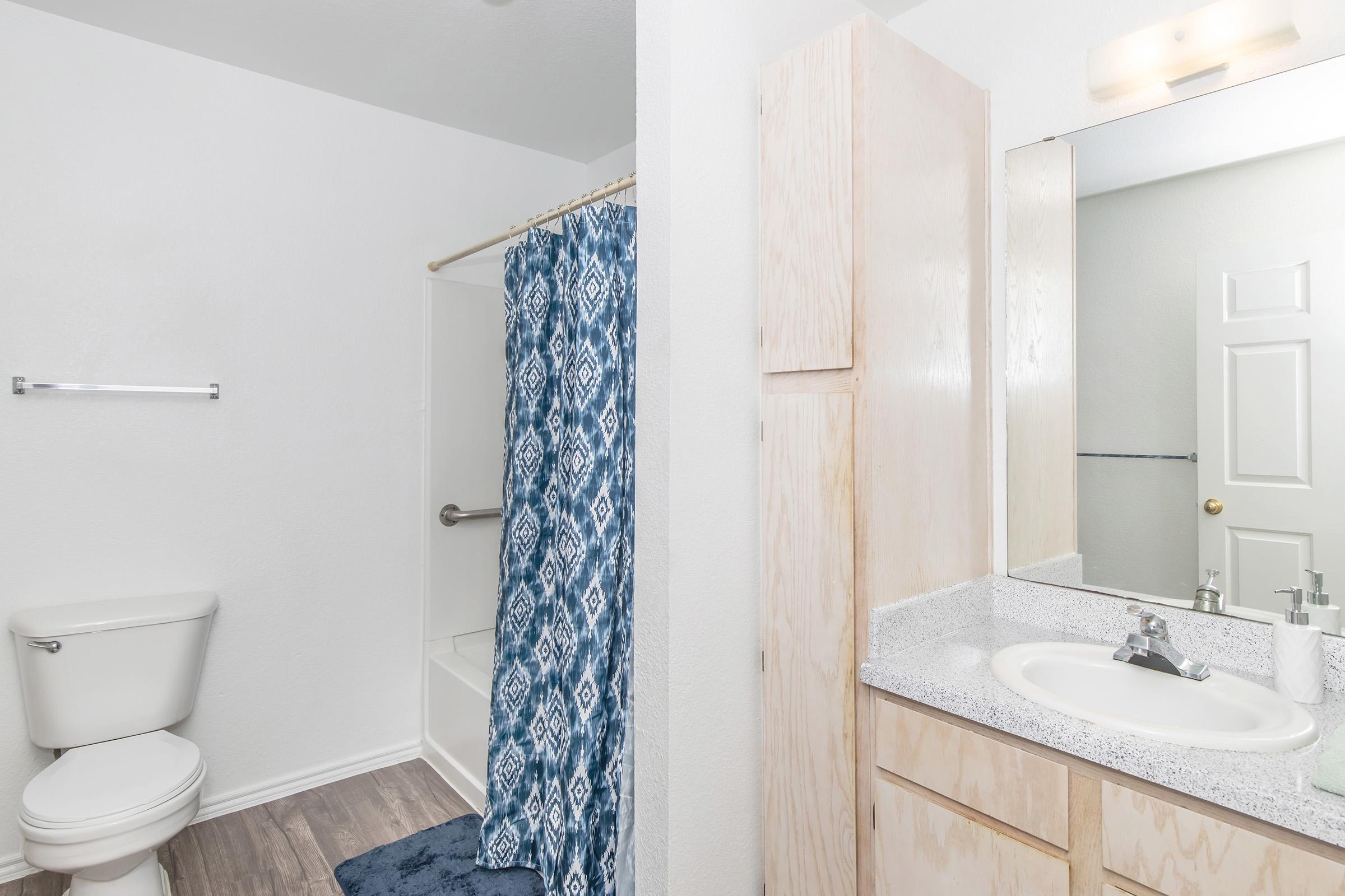 A clean bathroom featuring a toilet, a shower with a blue patterned curtain, and a sink with a countertop. Light-colored wooden cabinets are present, and the walls are painted white, creating a bright and spacious appearance.