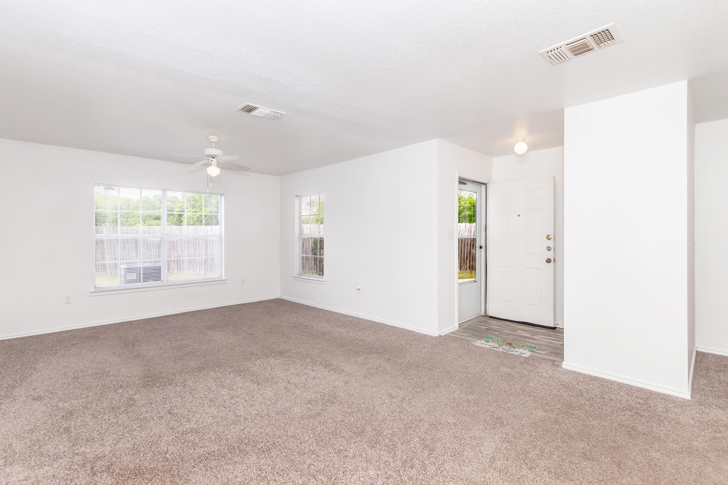 A spacious, empty living room with light-colored walls and carpet. The room features a ceiling fan and large windows allowing natural light. A front door is visible on the right, leading outside. The overall atmosphere is bright and inviting, ideal for personal customization.
