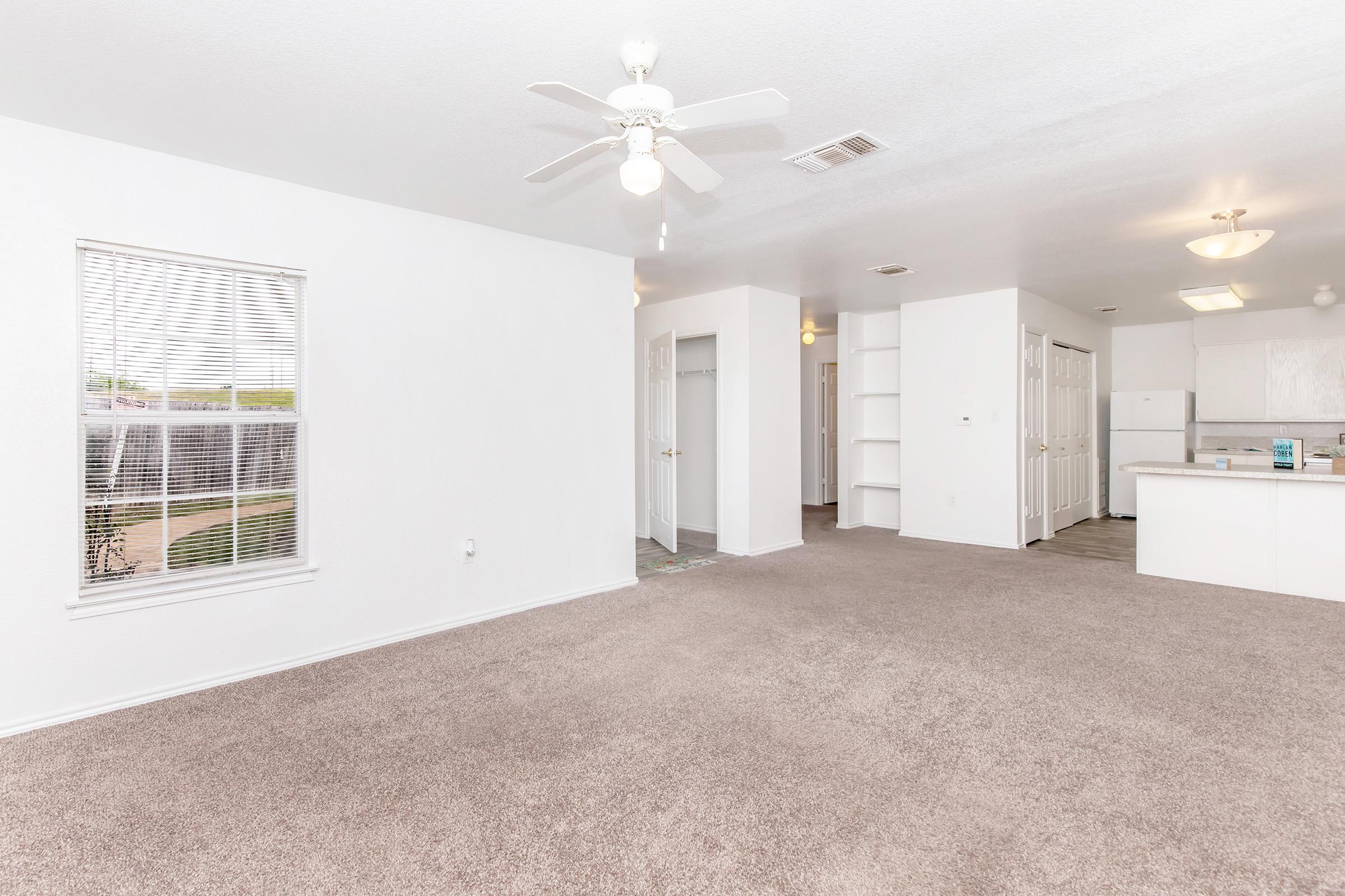 A spacious, well-lit living area featuring beige carpet, a ceiling fan, and a large window with blinds. The room leads to an open kitchen space with white cabinets and appliances. A hallway is visible, leading to additional rooms. The walls are painted white, enhancing the bright and airy atmosphere.