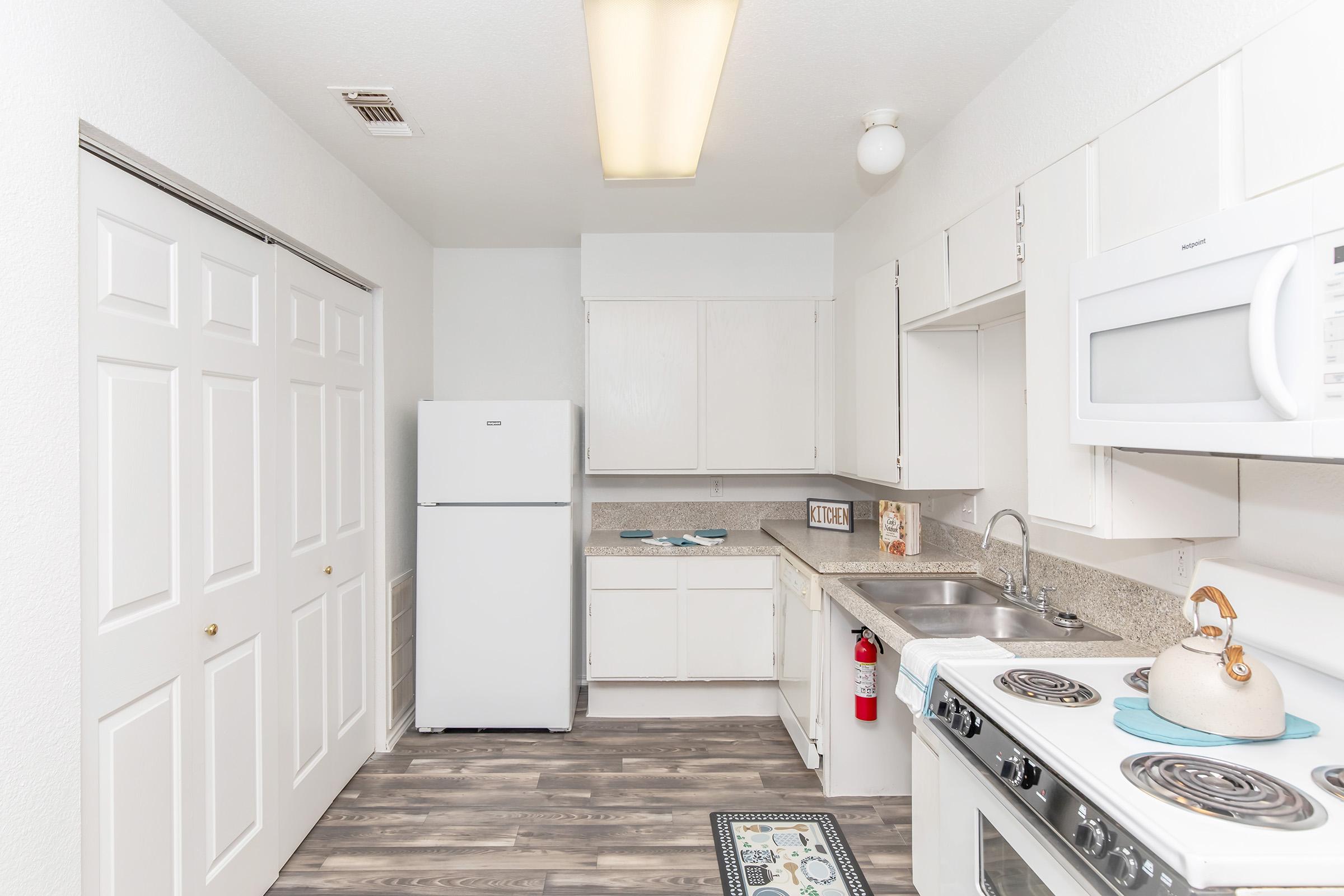 A modern kitchen featuring white cabinetry, a stainless steel sink, and a microwave above the stove. There is a refrigerator against one wall, and the floor has a wood-like finish. The kitchen is brightly lit with a ceiling light and includes a small decorative rug by the sink.