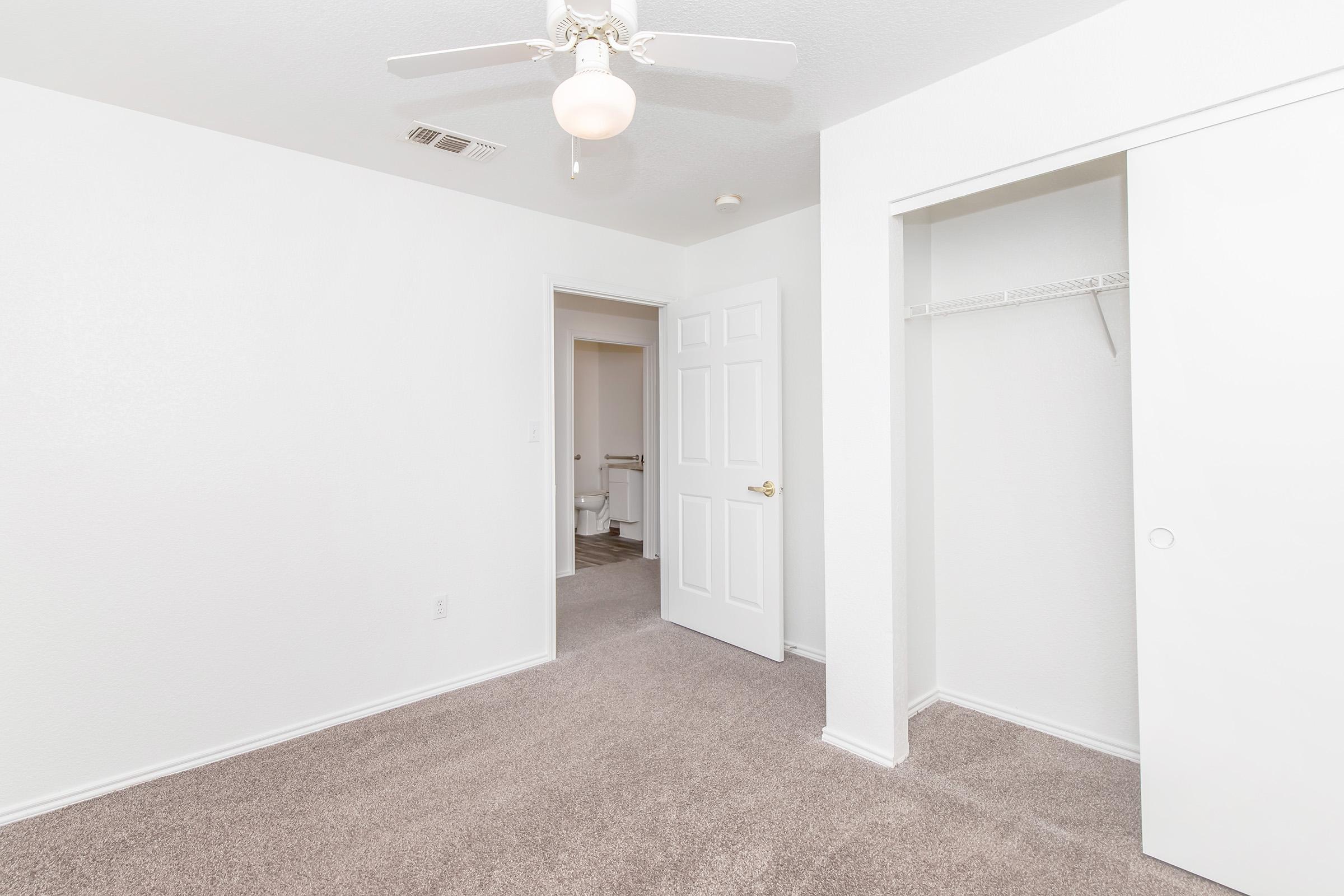 A bright, empty room with a ceiling fan, light beige carpet, and white walls. A closet with sliding doors is on the right, and an open door leads to a bathroom in the background. The space looks clean and inviting, suitable for various uses.