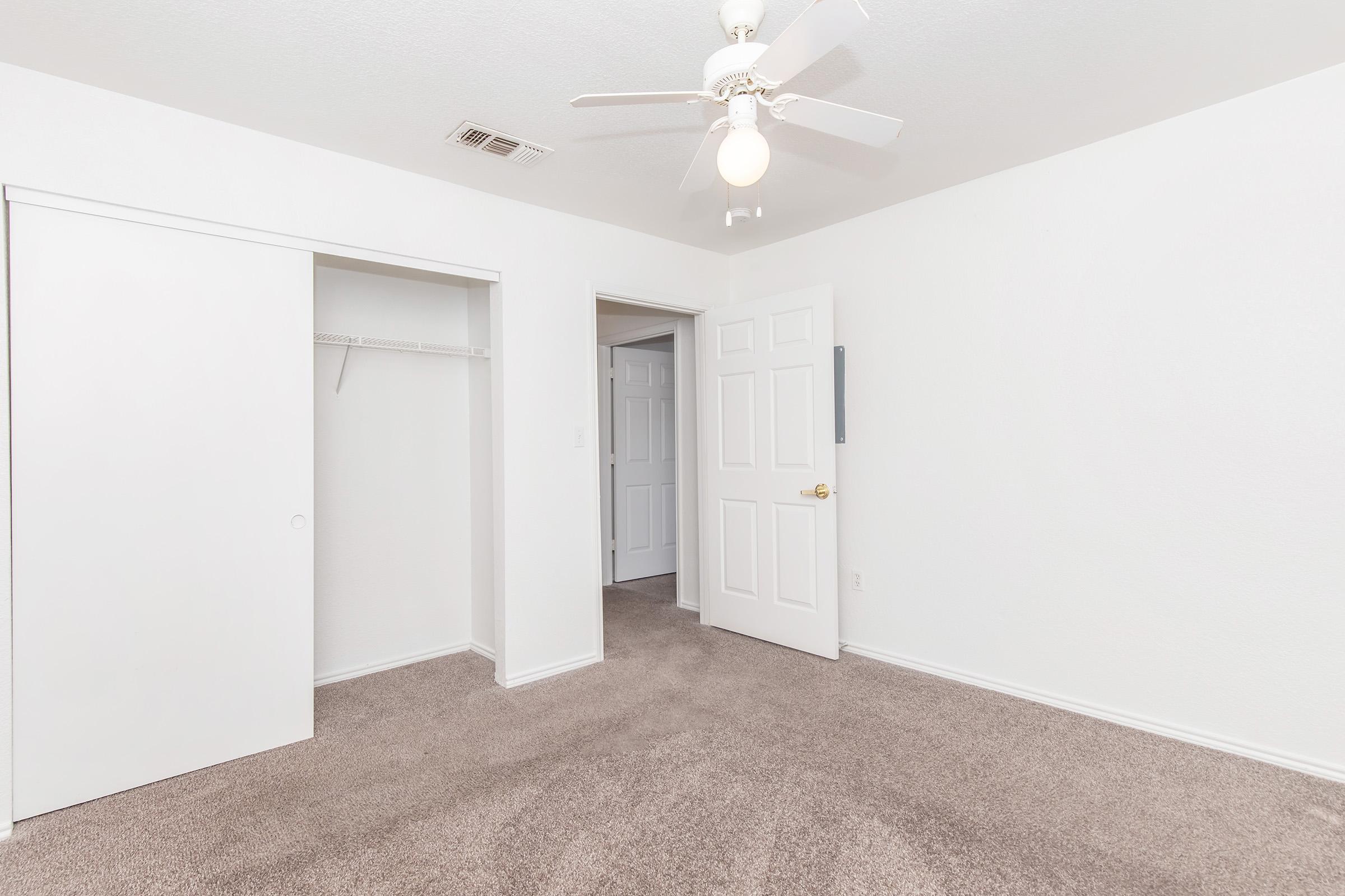 A clean, empty bedroom featuring light-colored walls and plush carpet. A ceiling fan hangs from the ceiling, and there is a closed door leading to another room. A closet with sliding doors is visible on one side, contributing to the spacious feel of the room.