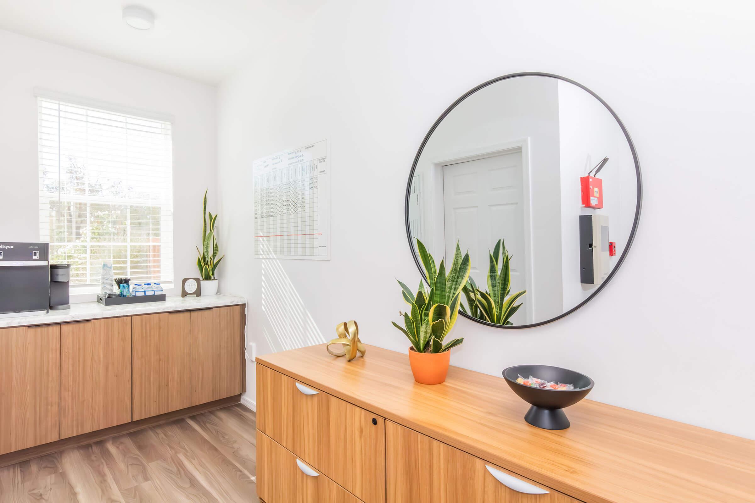 A modern office interior featuring a wooden storage unit, a round mirror on the wall, a potted snake plant, and a decorative bowl. There's a desk with office equipment and a whiteboard in the background. Natural light floods the space through a window, creating a bright and inviting atmosphere.