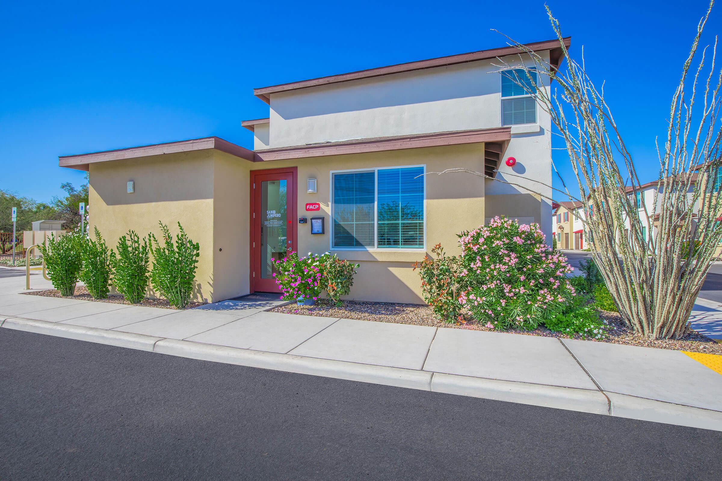 Two-story residential building with a beige exterior and a red door, surrounded by green shrubs and flowering plants. The clear blue sky is visible overhead, and a sidewalk runs in front of the property, indicating a neat and well-maintained area.