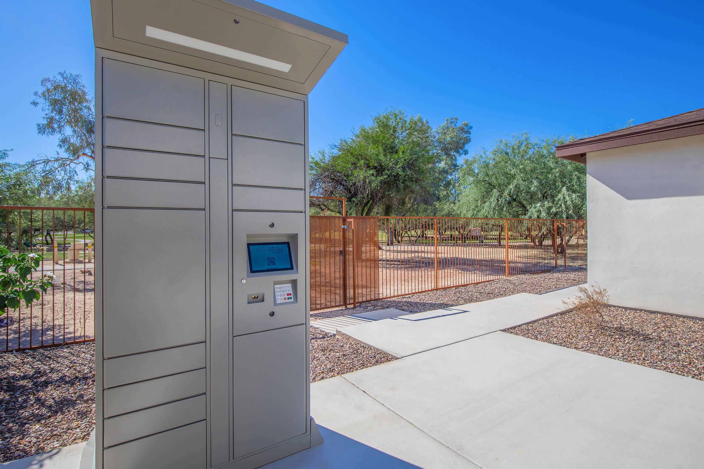 A modern, gray parcel locker system stands on a concrete pathway, with a touchscreen interface visible. In the background, there are trees and a fenced area, suggesting an outdoor setting. The sky is clear and blue, indicating a sunny day.